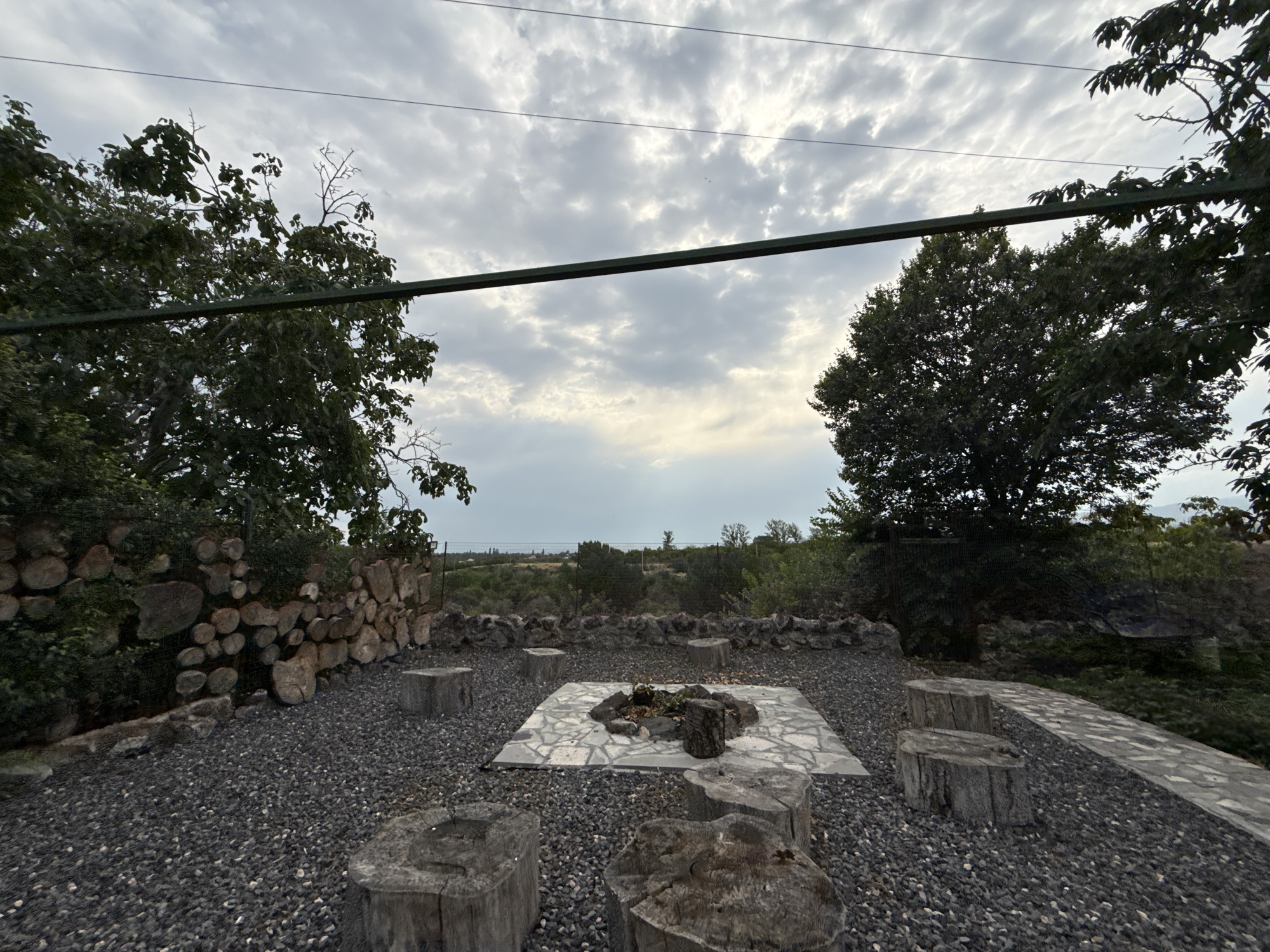 This image depicts an outdoor seating area centered around a fire pit, located in Ashtarak, Armenia. The fire pit is situated on a square flagstone platform within a gravel-covered courtyard. Several tree stump seats are arranged around the fire pit. To the left, a stone wall is partially covered by stacked logs and greenery. In the background, a fence separates the courtyard from a rural landscape with trees and rolling hills. The sky above is overcast with dramatic clouds. There are no people or animals visible in the scene. The overall atmosphere is rustic and natural.