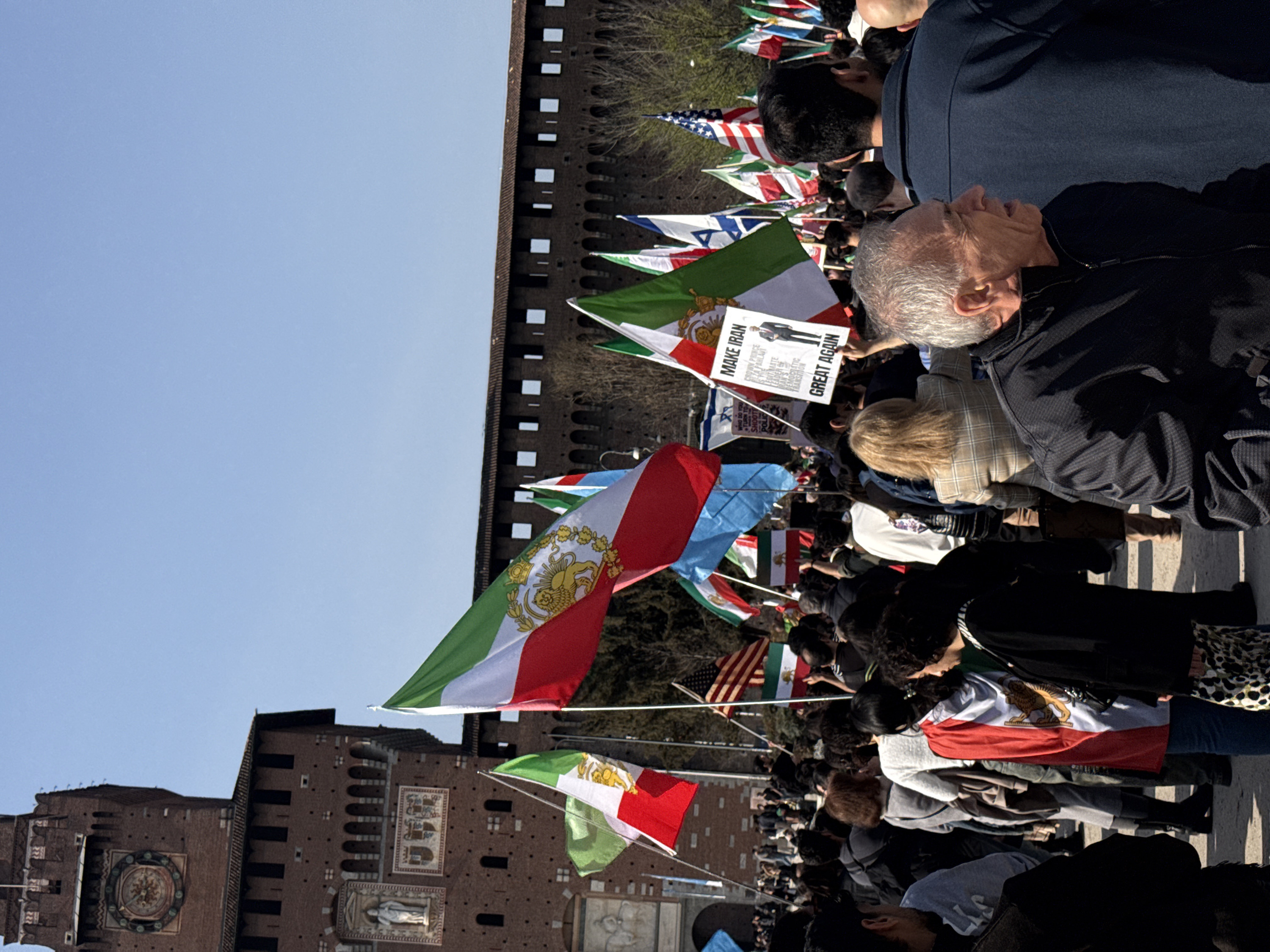 The image depicts a large outdoor gathering, identified as a demonstration, taking place in front of the Sforza Castle (Castello Sforzesco) in Milan, Italy. The castle's brown brick facade, featuring a prominent clock tower (Torre del Filarete) with an ornate clock face, battlements, arched windows, and decorative carvings, dominates the background under a clear blue sky. Bare trees are visible behind the castle's walls.
A dense crowd of individuals, numbering in the hundreds, occupies the middle and foreground. The participants appear diverse in age and gender and are dressed in casual attire, including jackets and sweaters. Many individuals are holding and waving flags. Predominant among these are numerous historical Iranian flags, characterized by green, white, and red horizontal stripes with the central gold Lion and Sun emblem. Other flags visible include at least one United States flag, one Israeli flag, and various solid-color or striped flags.
In the middle-right of the frame, a person holds a sign that reads "MAKE IRAN GREAT AGAIN" in large white letters. The sign also displays an image of a man in a suit and smaller text that includes phrases like "WHO DO YOU TURN TO WHEN THERE'S NO ONE LEFT TO CALL THE SHOTS? POLICE." and "REZA PAHLAVI IS THE LEGIMATE LEADER OF IRAN'S DEMOCRATIC TRANSITION GREAT AGAIN." Several individuals in the crowd have the historical Iranian flag draped over their shoulders or bodies.
In the immediate foreground, an older man with grey hair, wearing a black zippered jacket, is visible in profile on the right, looking towards the right side of the frame. Behind him, another man with dark hair and a dark blue jacket is seen from the back. The ground beneath the crowd appears to be a paved or solid outdoor surface.