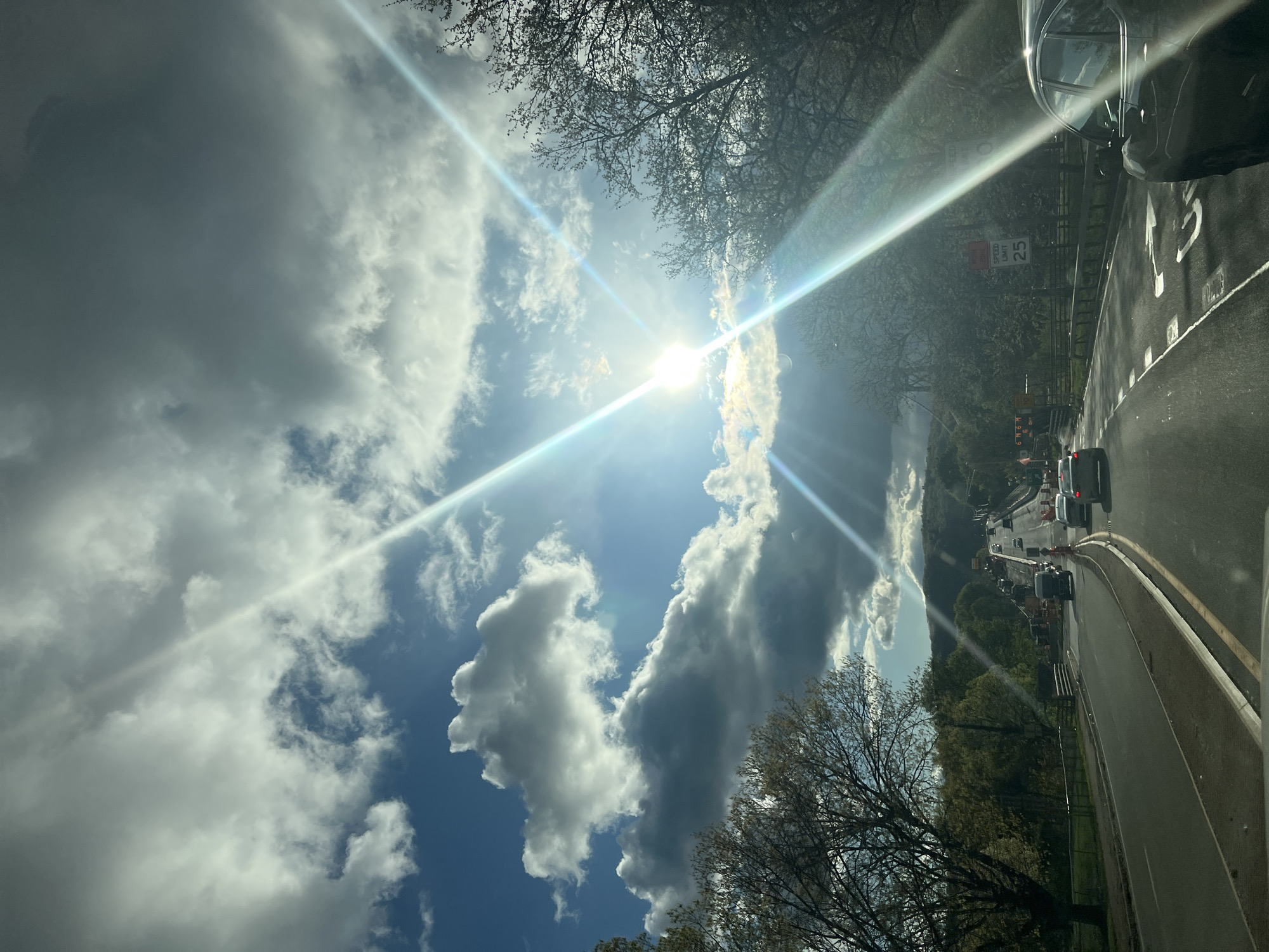This image captures a vibrant, sunlit scene from the perspective of a vehicle on a road, likely in the City of Poughkeepsie, United States of America. The dominant feature is a bright blue sky filled with a mix of fluffy white cumulus clouds and some darker, heavier grey clouds, suggesting variable weather. The sun is intensely bright, positioned in the upper left quadrant, and casts prominent, star-shaped lens flares across the image, indicative of a direct view towards it.
The right side of the scene depicts a multi-lane asphalt road with clear yellow double lines and white lane markings. Several vehicles are visible, appearing to be in traffic or moving slowly. A dark car's side mirror and portion of the roof are visible in the upper right, confirming the viewpoint is from inside another vehicle. Alongside the road, numerous trees with mostly bare branches suggest late winter or early spring. Further down the road, patches of green foliage are visible. Orange traffic cones and possibly roadwork equipment or temporary signs line the roadside, indicating active road construction or maintenance.
In the mid-ground, a white "SPEED LIMIT 25" sign is clearly visible on the right, providing a location cue for a regulated roadway. Below it, a smaller red sign is present but its text is unreadable. In the distance, on the left side, rolling hills or mountains covered with trees are visible, partially obscured by the cloud cover. The overall impression is one of a busy daytime drive under a bright, partly cloudy sky, with potential delays due to roadwork.