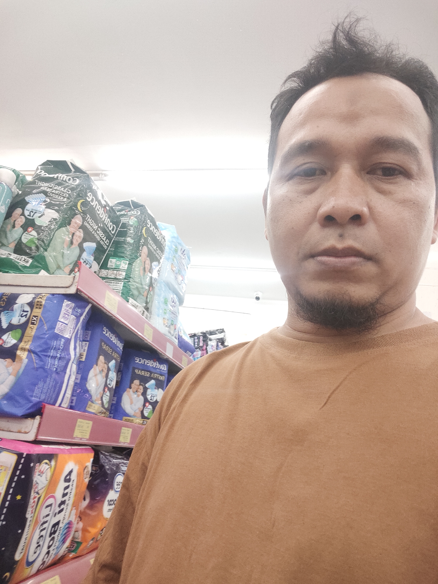 A medium-shot, front-facing selfie is captured from the chest up, featuring a man with short, dark, messy hair and a goatee. He is wearing a brown crew-neck t-shirt and stands in an aisle of a retail store. Behind him, shelves are stocked with various packaged goods. Prominently displayed on the shelves are large packages of adult diapers, some in blue packaging with images of elderly couples and the text "Confidence" and "Extra Serap". Other packages are green and also appear to be diapers. The lighting suggests an indoor, well-lit environment, consistent with a supermarket or pharmacy. The man's facial expression is neutral, and he is looking directly at the camera. The background merchandise is out of focus, drawing attention to the subject. The scene is likely within the commercial sector of Desa Banyumas, Indonesia, as suggested by the presence of retail goods and the likely demographic of the individual.