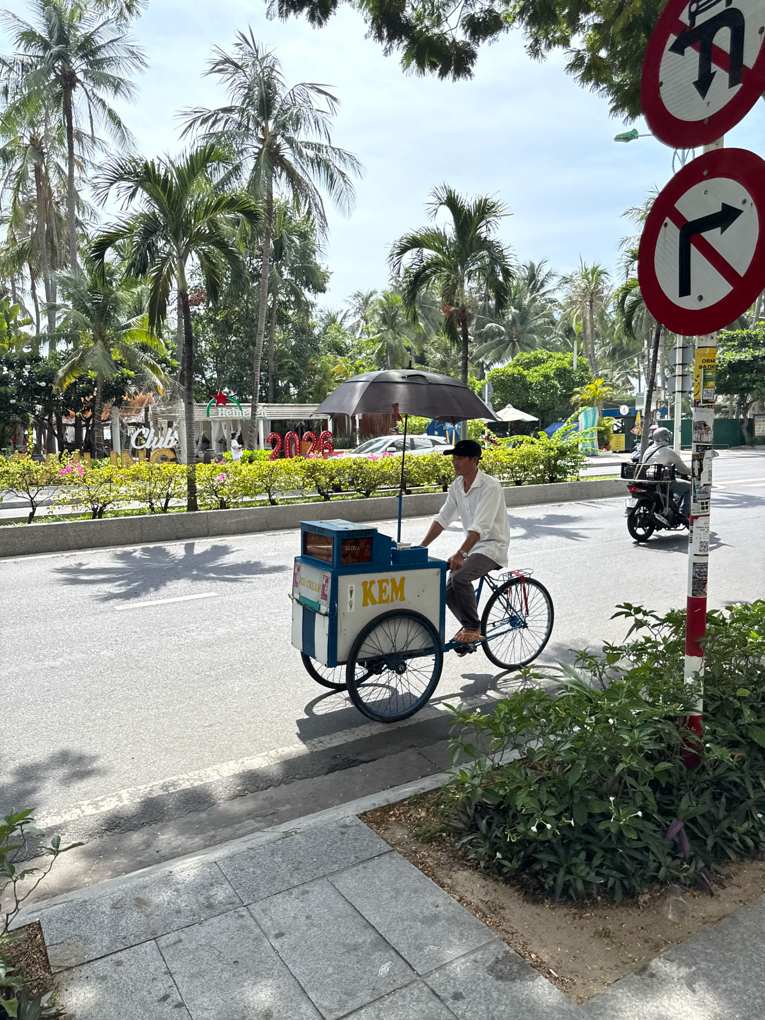 A man wearing a white shirt, dark pants, and a black cap rides a blue and white ice cream cart with an umbrella on a sunny day in Nha Trang, Vietnam. The ice cream cart has "KEM" written on its side. In the background, palm trees, lush green bushes, and a building with "Club's" and "2026" are visible. Traffic signs indicating no U-turns and no right turns are posted on a pole. A motorcycle is also seen on the road. The scene is set on a paved street with a sidewalk.