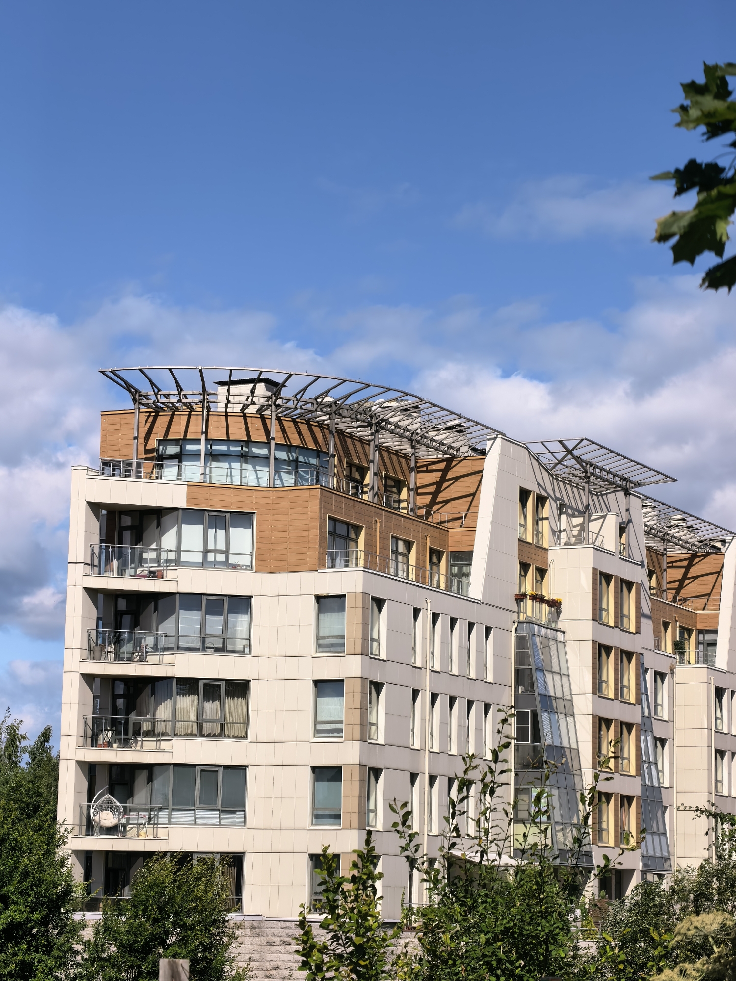 This is a vertical, eye-level, medium shot of a modern apartment building in Sestroretsk, Russia. The building has a complex, multi-faceted design with light beige and brown facade panels. It features numerous windows and balconies, many with glass railings. A distinctive feature is the curved, metallic roof structure with intricate metal supports extending over the upper floors. In the foreground, lush green trees and foliage partially obscure the lower levels of the building. The sky above is a clear blue with scattered white clouds. There are no people visible in the image.