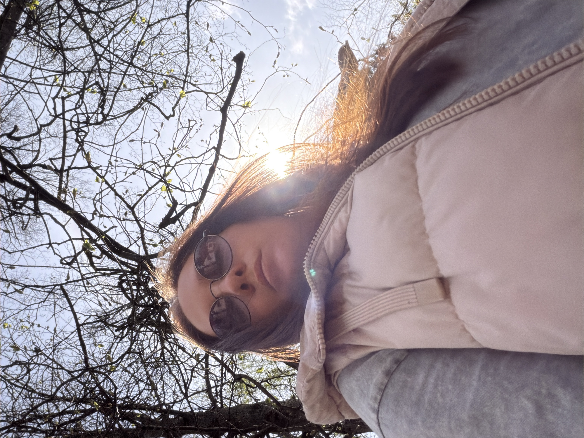 A low-angle shot shows a woman in sunglasses looking down at the camera, with leafless tree branches filling the background sky. She is wearing a light pink puffer vest over a gray top. The sun is visible behind her head, creating a glare. The location is outdoors, implied to be Tokkari, Russia by external context.