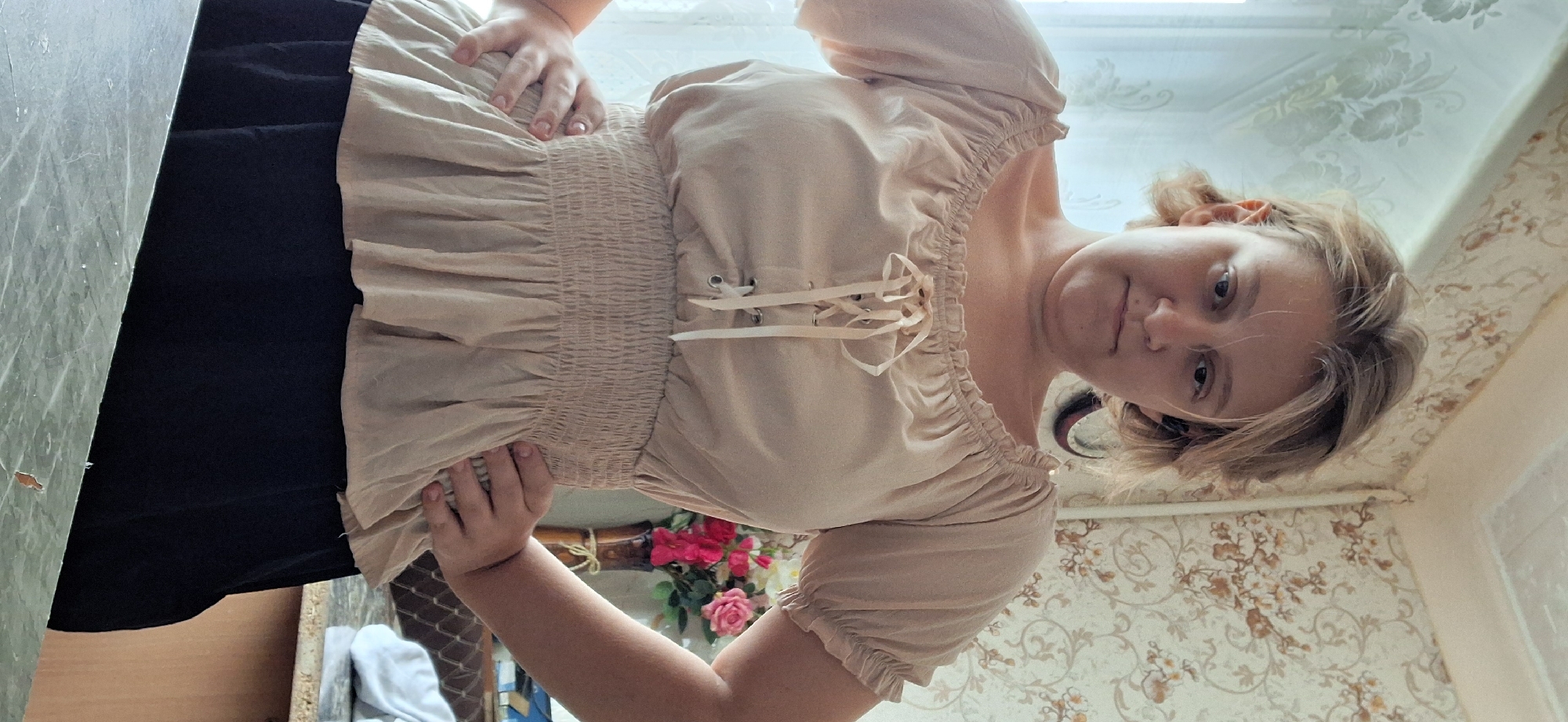 A young woman stands indoors, facing the camera with her hands on her hips. She has short, light brown hair and is wearing a light-colored, off-the-shoulder blouse with a cinched waist and a black skirt. The background shows a patterned wallpaper, a window with a curtain, and a vase of flowers on a table. The scene appears to be a residential interior, possibly in the city of Petropavl, Kazakhstan.
