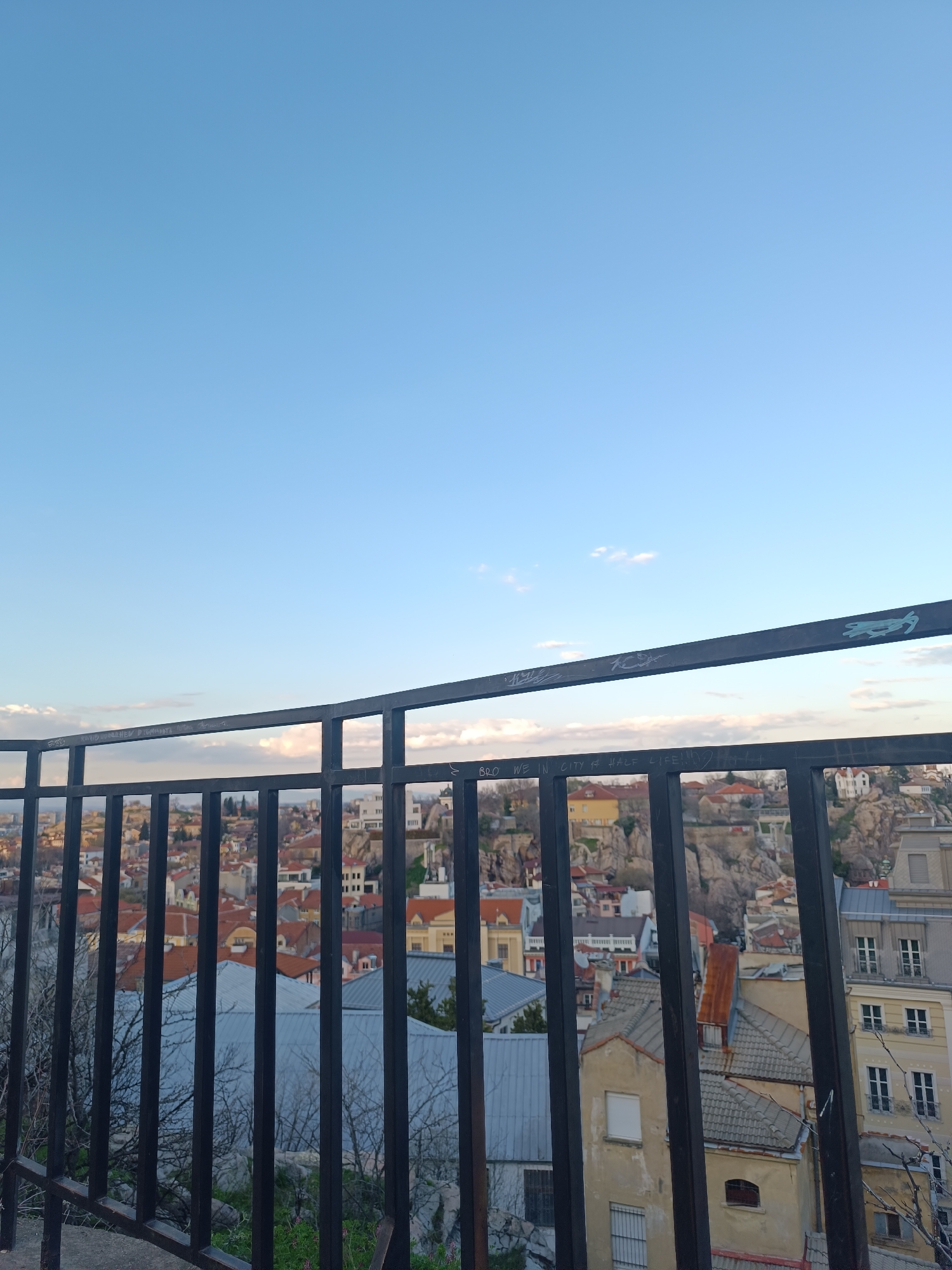 The image captures a panoramic view of Plovdiv, Bulgaria, seen through a black metal railing. The foreground is dominated by the railing, partially obscuring the cityscape. Behind the railing, a dense arrangement of buildings with red and gray roofs fills the middle ground. Rocky outcrops are scattered among the buildings. In the background, a pale blue sky fades into the horizon. The image is taken during daylight hours, indicated by the lighting conditions. The perspective suggests that the photograph was taken from an elevated vantage point.