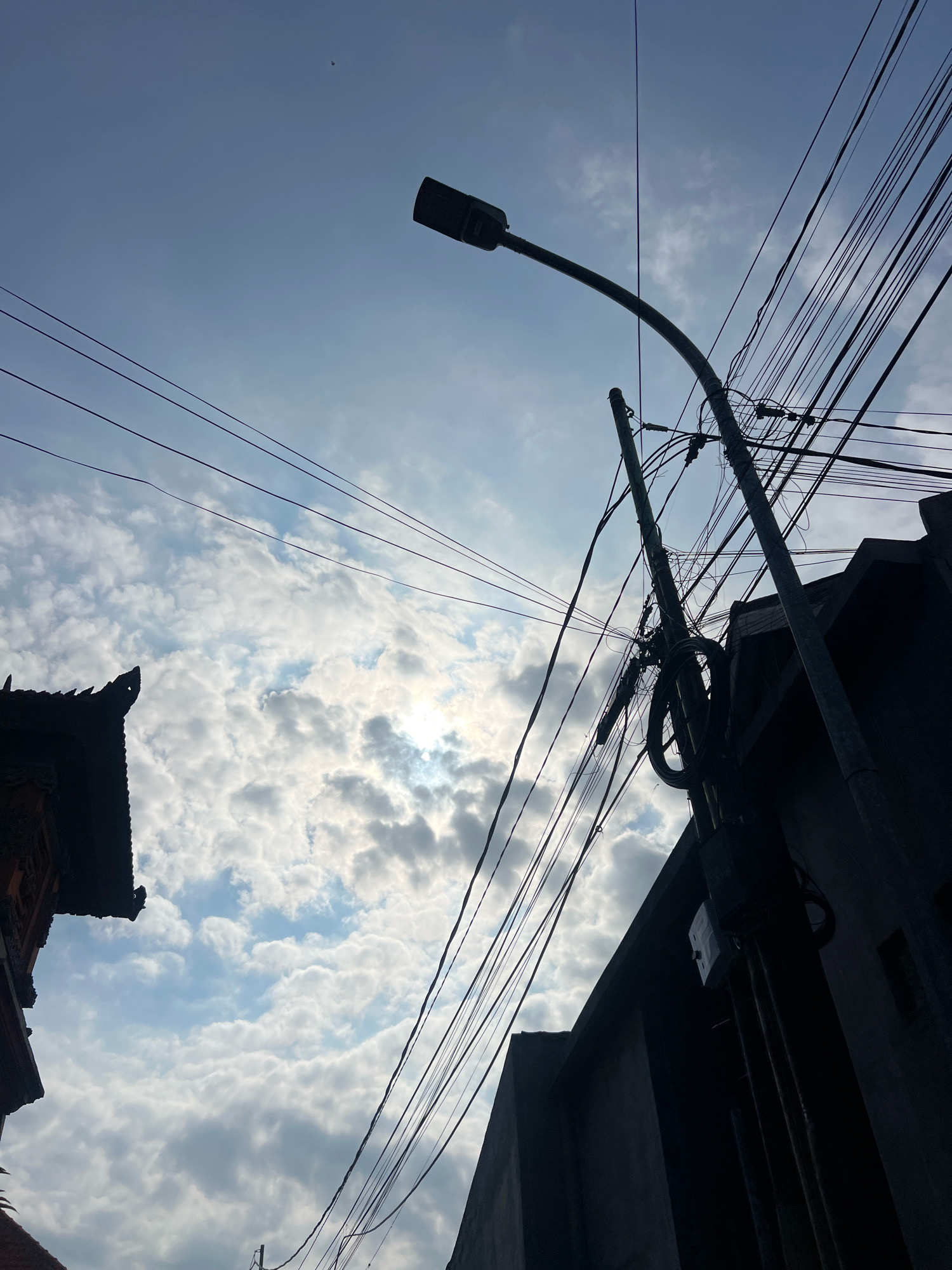 An upward shot captures a utility pole with numerous electrical wires against a cloudy sky in Kuta, Indonesia. The sky is a mixture of blue and white clouds, with sunlight peeking through in some areas. The utility pole is tall and dark, with a street lamp attached to it. Several black wires are tangled around the pole and stretch across the frame. To the left, a silhouette of a building with a decorative roofline is visible. To the right, a dark, angular building with a rectangular window is partially seen. The image has a high contrast, with the subjects silhouetted against the bright sky. There are no people or animals present in the image.