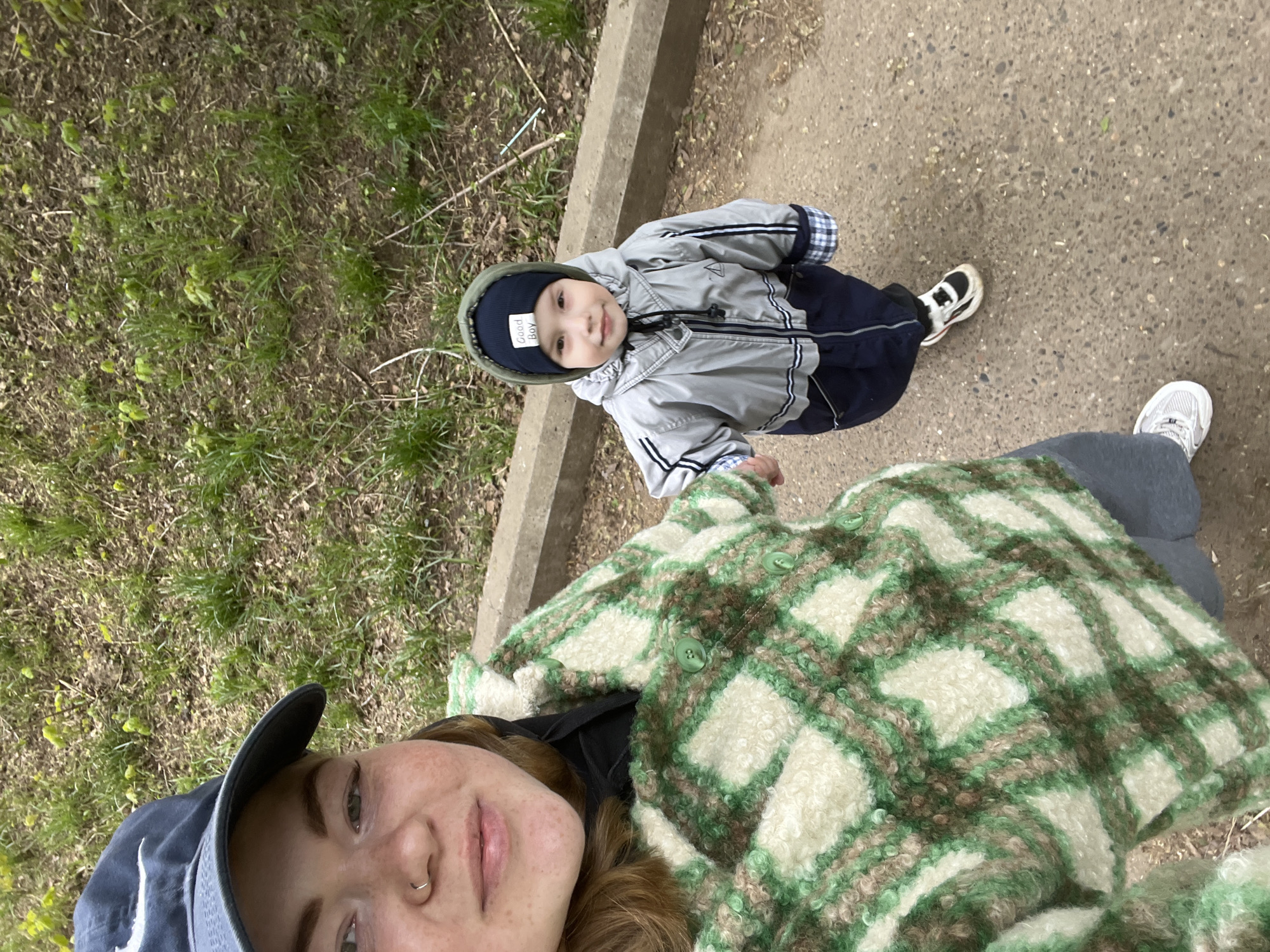 A selfie taken outdoors shows a woman and a small child. The woman, wearing a blue baseball cap and a green and white plaid jacket, holds the camera. The child, wearing a gray jacket, navy pants, and a navy hat labeled "Good Boy", stands to her right. The setting appears to be a paved pathway with grass and greenery to the side. A concrete curb is visible near the grass. There is no information in the image that would indicate it was taken in the city of Chaikovsky, Russia.