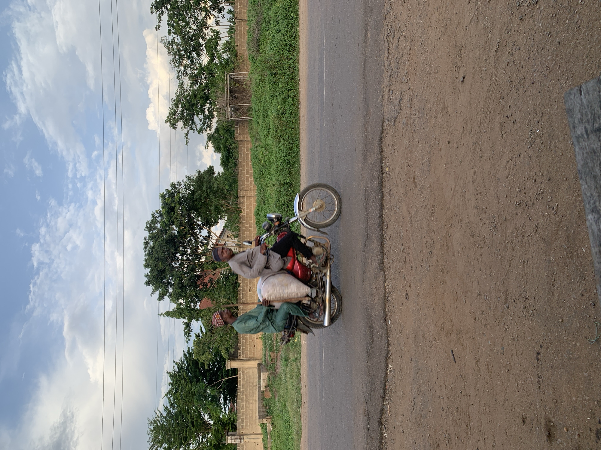 The image depicts two adult males riding a red motorcycle on a paved road in Gbeleko, Nigeria, under a sky with scattered clouds. The driver, wearing a grey traditional garment and a dark cap, operates the motorcycle, while the passenger, dressed in a green traditional garment and a patterned cap, sits behind him. A large, off-white, cylindrical sack is positioned on the motorcycle between the driver and the passenger, with the passenger appearing to stabilize it. A long, thin stick extends upwards from behind the passenger.
The motorcycle has chrome accents and spoked wheels. To the immediate left of the paved road is an unpaved, reddish-brown dirt shoulder. The background features a brick or concrete wall topped with lighter coping, obscured in parts by dense green foliage and trees. Beyond the wall, indistinct building structures are partially visible. Further to the right, behind a dense patch of green shrubbery, a weathered wooden structure, possibly a shelter or goalpost, is present. Multiple dark power lines traverse the upper portion of the sky. The lighting indicates natural daylight, likely diffused.