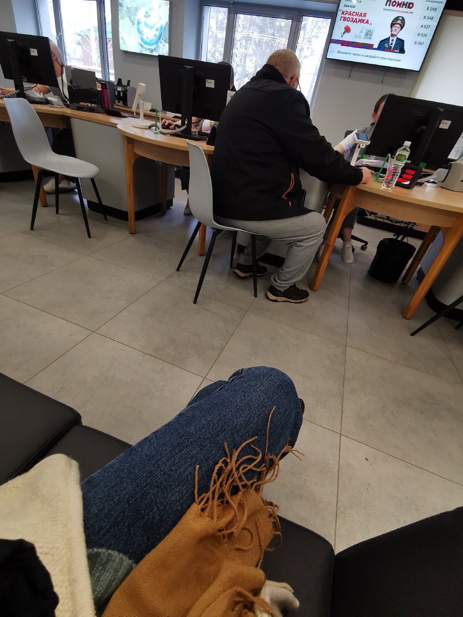 The image captures the interior of a government office, likely in Russia, as suggested by the signage in Russian. The scene is composed from a first-person perspective, focusing on a pair of legs clad in denim jeans, overlaid with a tan-colored textile item, possibly a scarf or wrap. The legs rest on a black leather chair or sofa.
In the background, an office environment unfolds. Several individuals are present: one man in a black jacket and grey sweatpants is seated at a desk, appearing to interact with a computer. Another person is visible, partially obscured by the desk and computer equipment.
The office layout features a series of desks arranged in a row, each equipped with a computer monitor. One of the desks is occupied with office supplies, including a water bottle and a small electronic device with a digital display. Above the desk, a television screen displays a public service announcement, bearing an image of a man in a military uniform and the title “Pomno.”
The floor is tiled, and daylight streams in through several large windows, suggesting a bright and airy space. The overall impression is one of a typical government office, possibly a service center for citizens to receive assistance or conduct official business.