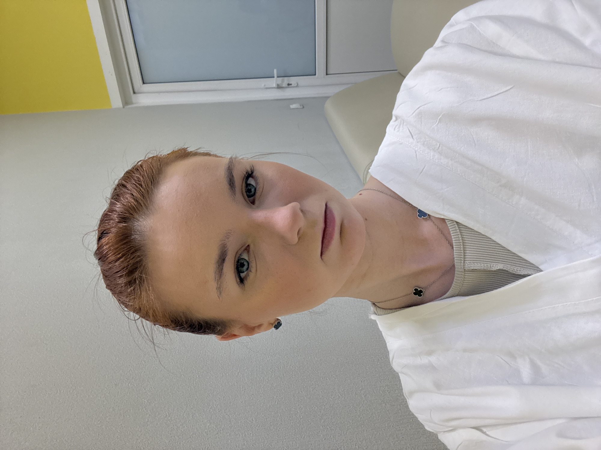 A fair-skinned woman with auburn hair is oriented slightly away from the camera. She is wearing a white shirt over a beige striped garment, and a necklace featuring a black clover-shaped pendant. She is in a room with light grey walls, a yellow upper wall section, and a window. A light-colored padded surface is visible in the background. The location is Unknown, Russia.