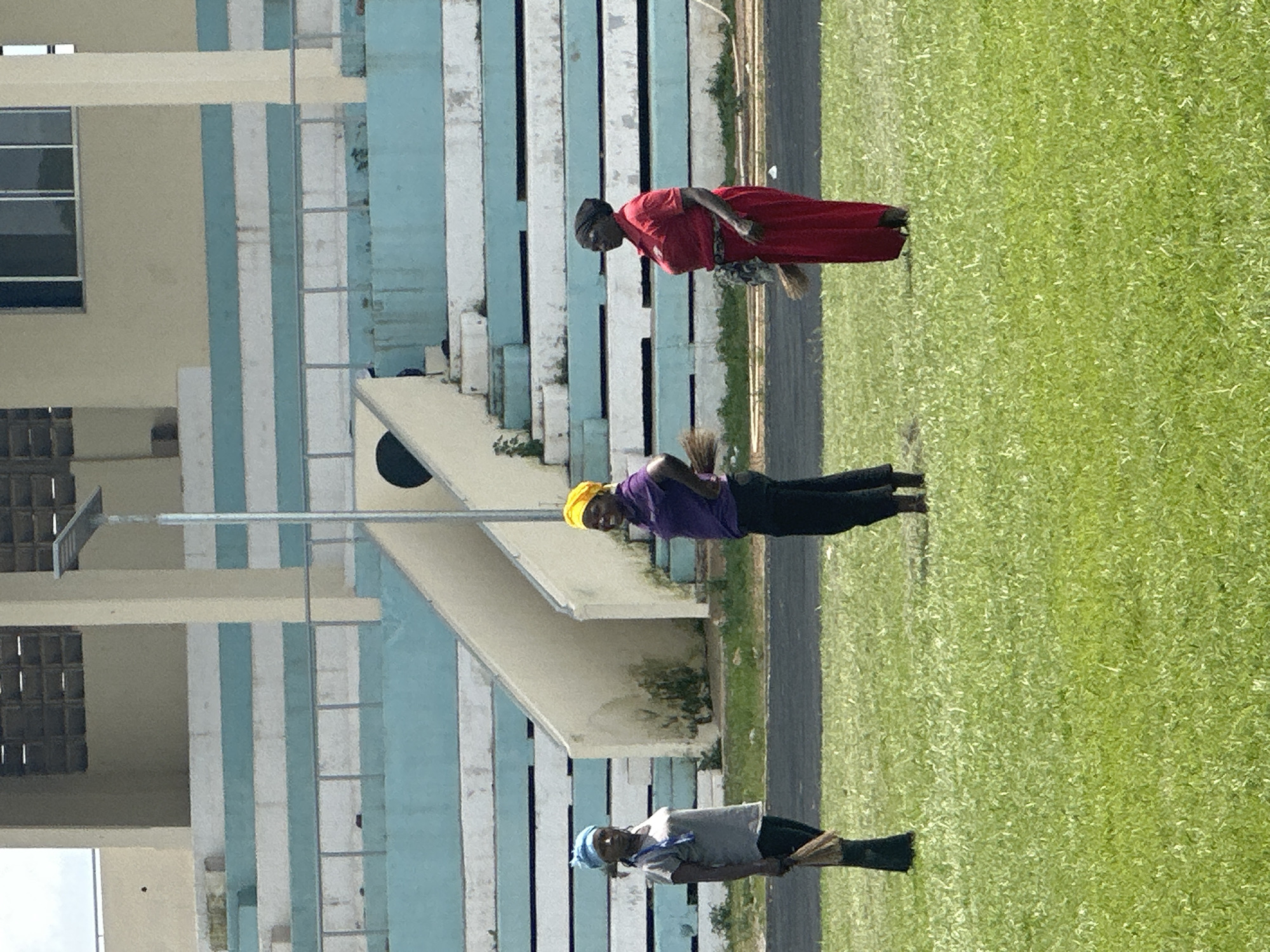 In Yelwa, Nigeria, three women stand on a grassy field in front of a stadium. The woman on the left wears a grey t-shirt, black pants, and a blue headscarf. She holds a broom in her right hand. The woman in the center wears a purple polo shirt, black pants, and a yellow headscarf, and also holds a broom. The woman on the right is dressed in a red dress, black pants, and a black headscarf, and she holds a broom. The stadium behind them features blue and white striped seating and a concrete structure. The sky is overcast.