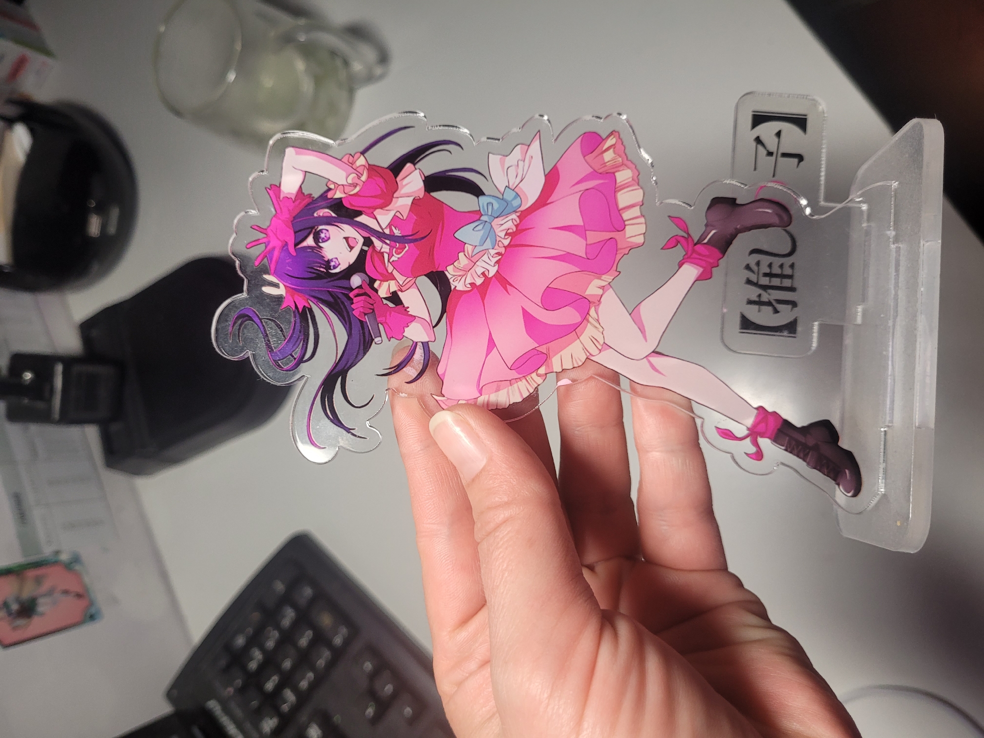 An adult's hand holds up an acrylic standee featuring an anime-style female character. The character has long, flowing dark purple hair, wide purple eyes, and a cheerful, open-mouthed expression. She is depicted in a dynamic, leaning pose, wearing a vibrant pink ruffled dress with a light blue bow, pink fingerless gloves, and dark boots with pink ribbon accents. She holds a silver microphone in her left hand. The standee has a glossy, reflective surface, likely due to being acrylic.
The standee is inserted into a clear acrylic base, visible on the right. This base prominently displays vertically oriented Japanese text: 【推し】 (Oshi) and a character above it, likely 【子】 (Ko), forming "Oshi no Ko," which is the title of a popular anime and manga series.
The background, largely out of focus, suggests an indoor setting, possibly a desk or workstation. Visible blurry objects include a clear glass or plastic container in the top left, a black rounded object (possibly a device or part of one) to the left, and parts of a black keyboard along with a small, colorful card or sticker featuring another anime character in the bottom left. The lighting is soft and appears to be artificial or diffuse natural light. The scene is located in Izhevsk, Russia.