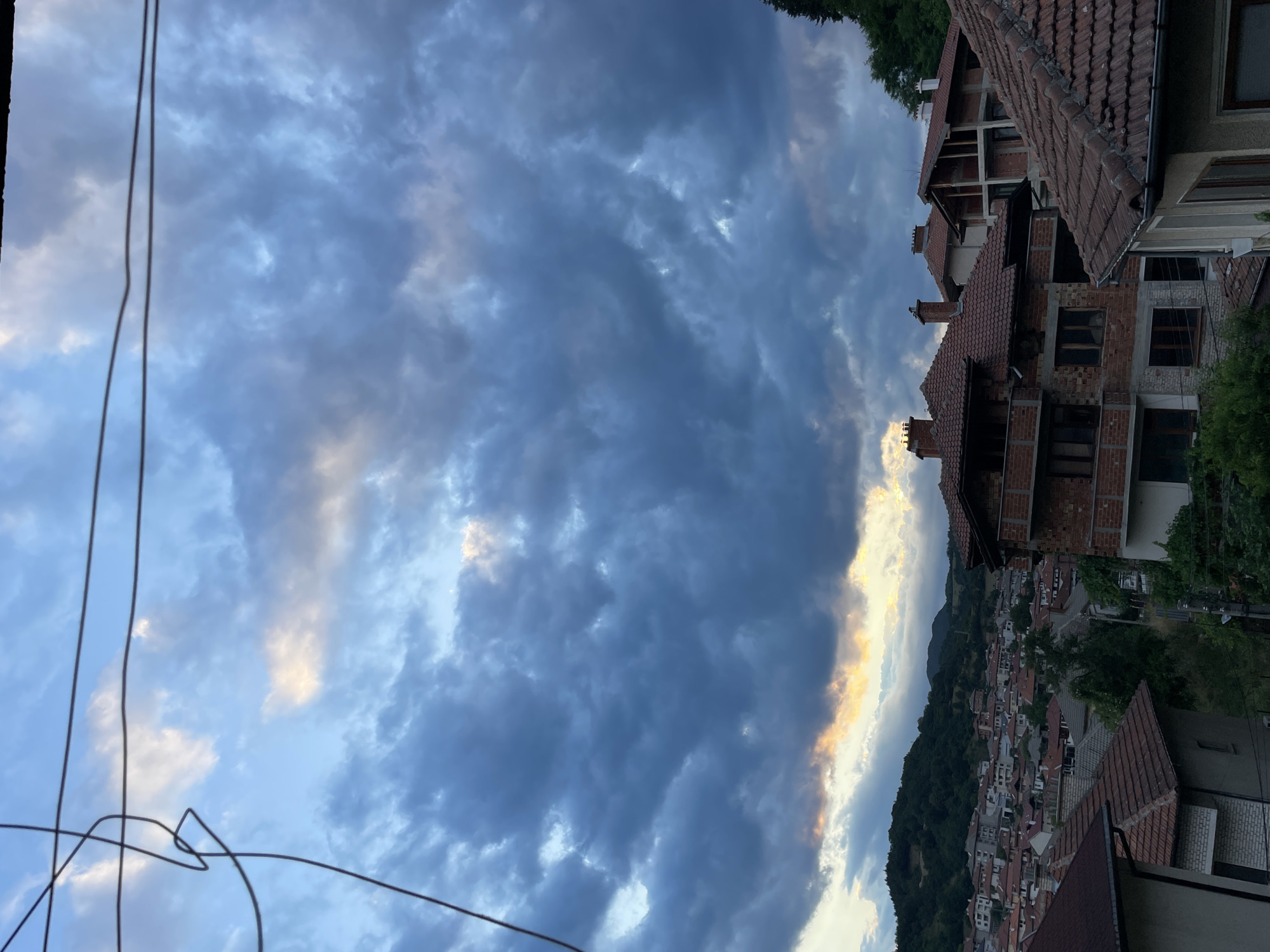 The image depicts a cloudy sky over a residential area in the city of Zlatograd, Bulgaria. The sky is filled with dark, dramatic clouds with patches of light breaking through near the horizon, suggesting either dawn or dusk. Below the sky, a cluster of houses with red tile roofs dominates the mid-ground. Some of these houses appear to be under construction, with exposed brickwork and unfinished elements. Trees and a hillside covered in vegetation form the backdrop to the urban landscape. Electrical wires are visible in the foreground and upper portion of the image. There are no people or animals visible in the scene. The overall atmosphere is one of a developing town under a dynamic sky.