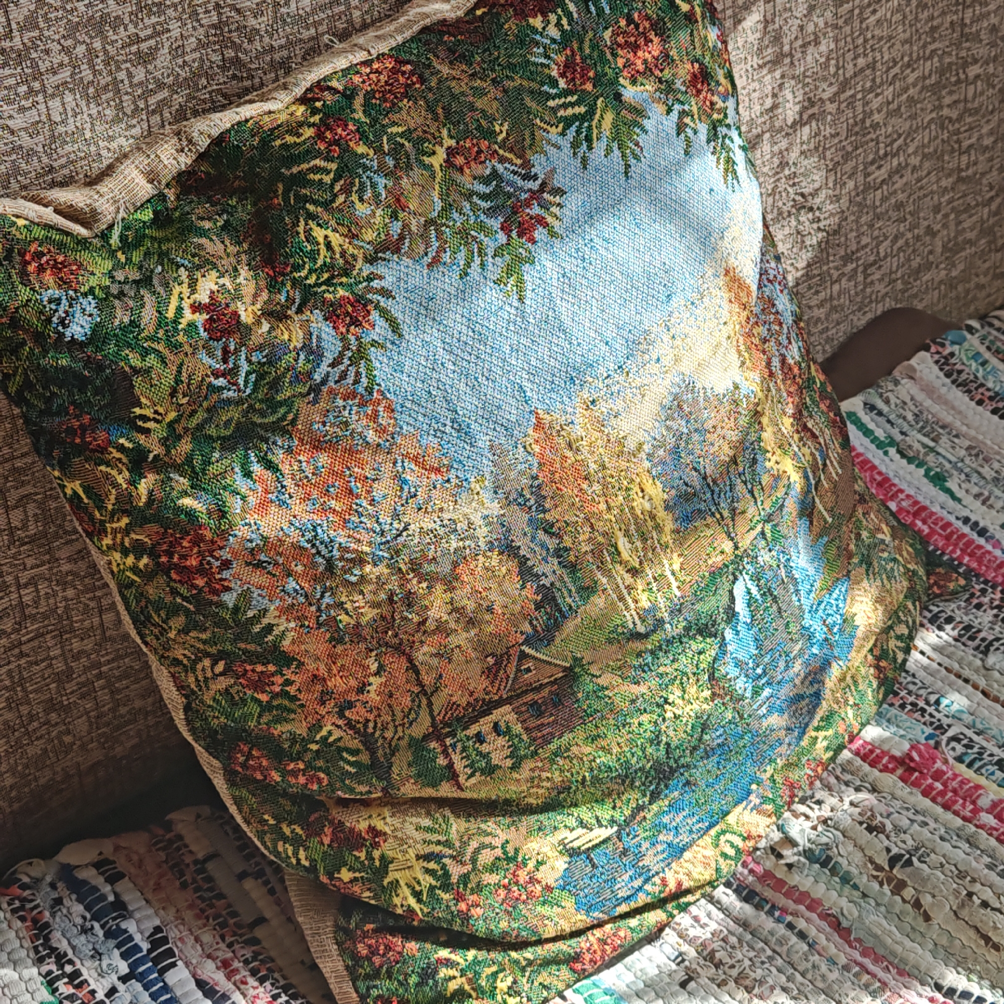 A close-up shot depicts a decorative pillow with a woven landscape pattern, resting against a couch with a beige fabric texture. The pillow features a sky-blue backdrop with details resembling trees and a cabin in the lower portion of the landscape. The scene is illuminated by sunlight that streams in from the side. The pillow is on a multicolored woven rag rug. The location is not discernible as Rostov, Russia, as there are no identifying indicators in the image.
