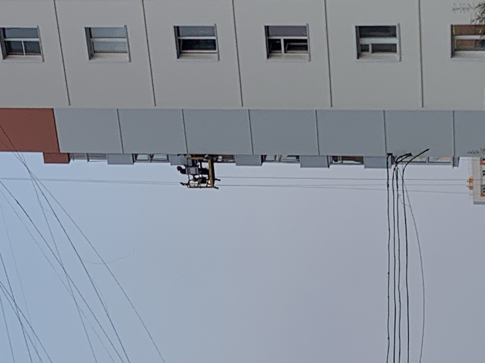 The image depicts a high-rise residential building in Norilsk, Russia, with a suspended platform carrying two individuals working on the exterior. The building features a light gray facade with multiple windows and a distinct red-orange section at the top. The individuals are positioned on a yellow, open-sided platform suspended by cables against the building's side. The surrounding environment is dominated by a pale, overcast sky, and a network of overhead wires cuts across the upper portion of the frame, suggesting an urban setting. The scene appears to capture ongoing exterior maintenance or construction work on the building.