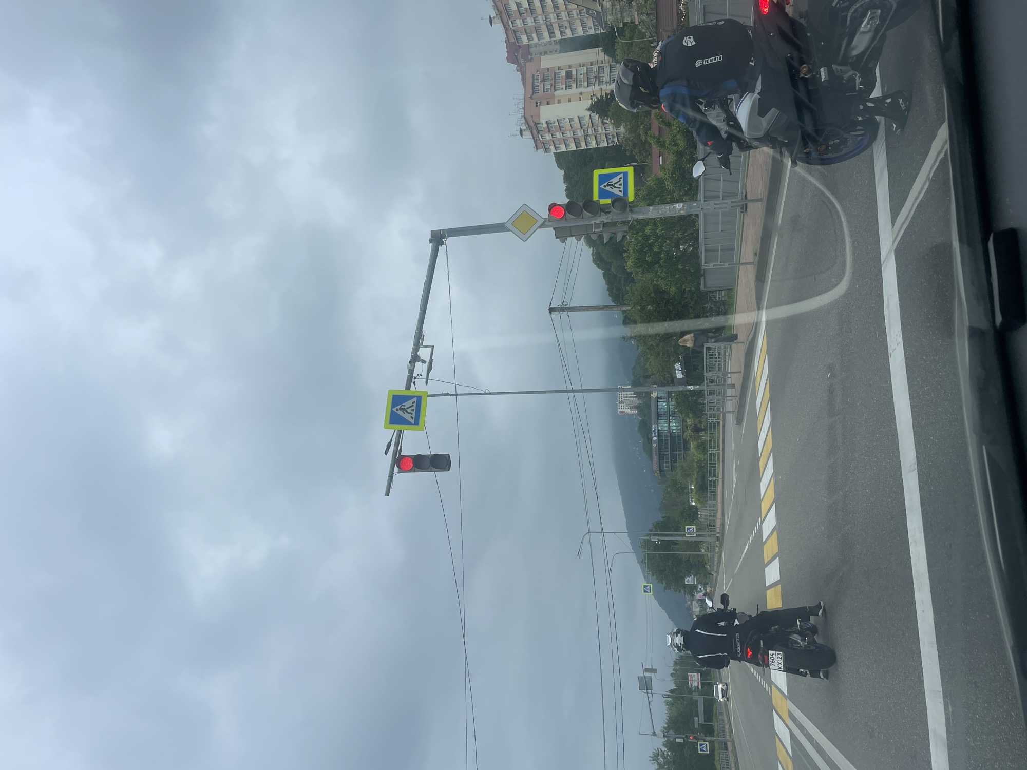 The photograph captures a street scene in Sochi, Russia, under an overcast sky. The view appears to be taken from inside a vehicle, as a portion of the window frame is visible on the right. The street is lined with crosswalks, each marked with zebra stripes and pedestrian crossing signs. A series of traffic lights is visible, all displaying red signals. Two motorcyclists are present: one is positioned near the top right corner of the image, and another is further down the road, near the center. Buildings are visible in the background, with mountains in the distance. Power lines stretch across the sky above the street.