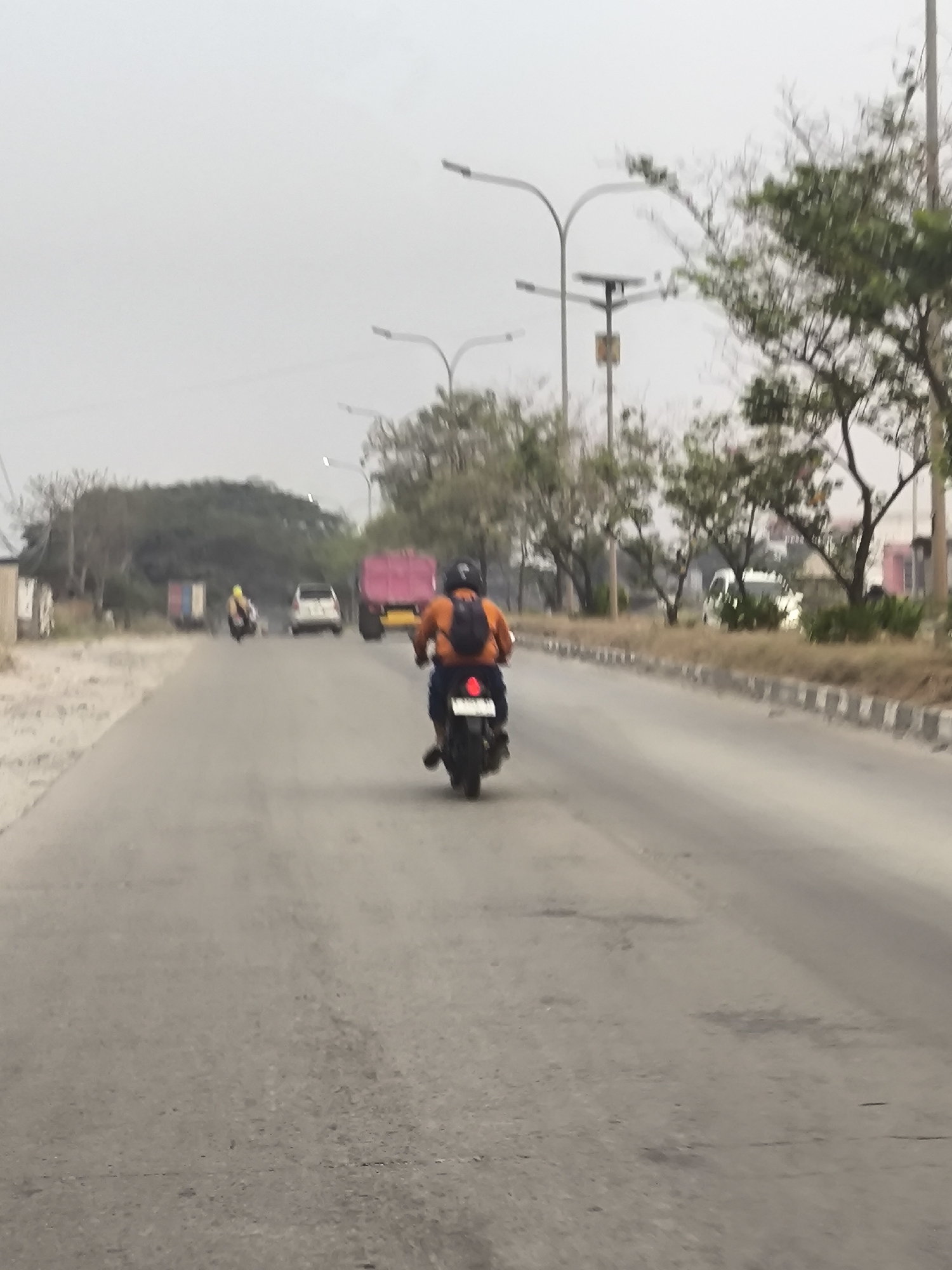 The image depicts a street scene in Cilegon, Indonesia. A person is riding a motorcycle in the foreground, wearing an orange shirt and a backpack. The motorcycle has a visible taillight. In the background, other vehicles are present, including a white car, a pink truck, and another motorcycle with a rider wearing a yellow helmet. The road is paved and appears to be a multi-lane highway. Lampposts are visible along the side of the road, some with solar panels. Trees and vegetation line the edges of the road, and there are some buildings and structures in the distance. The sky is overcast. The scene appears to be an outdoor, daytime environment.