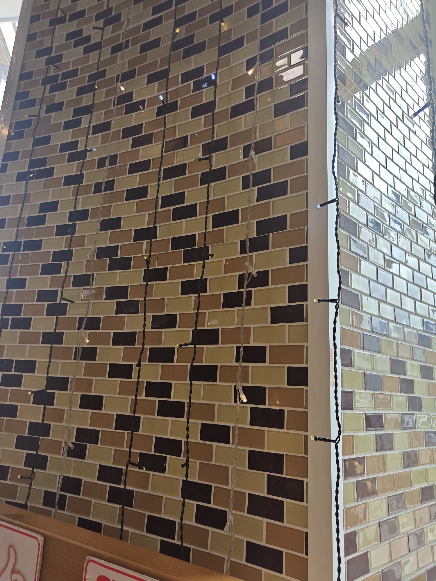 The image shows a close-up of a tiled wall with strings of lights. The tiles are rectangular and arranged in a brick pattern. The wall is divided into two sections by a white vertical beam. The left section features tiles in shades of brown, beige, and dark blue. The right section, which appears to be a corner, has tiles in lighter shades of green, beige, and off-white, with some purple-toned tiles mixed in. Thin black and white wires with small LED lights are draped vertically across the tiles. The lighting suggests an indoor setting, possibly a restaurant or shop. There are also glimpses of wooden surfaces and signs at the bottom of the frame, indicating a commercial space. The image does not provide any specific landmarks or clues to pinpoint its location within the city of Petropavl, Kazakhstan.