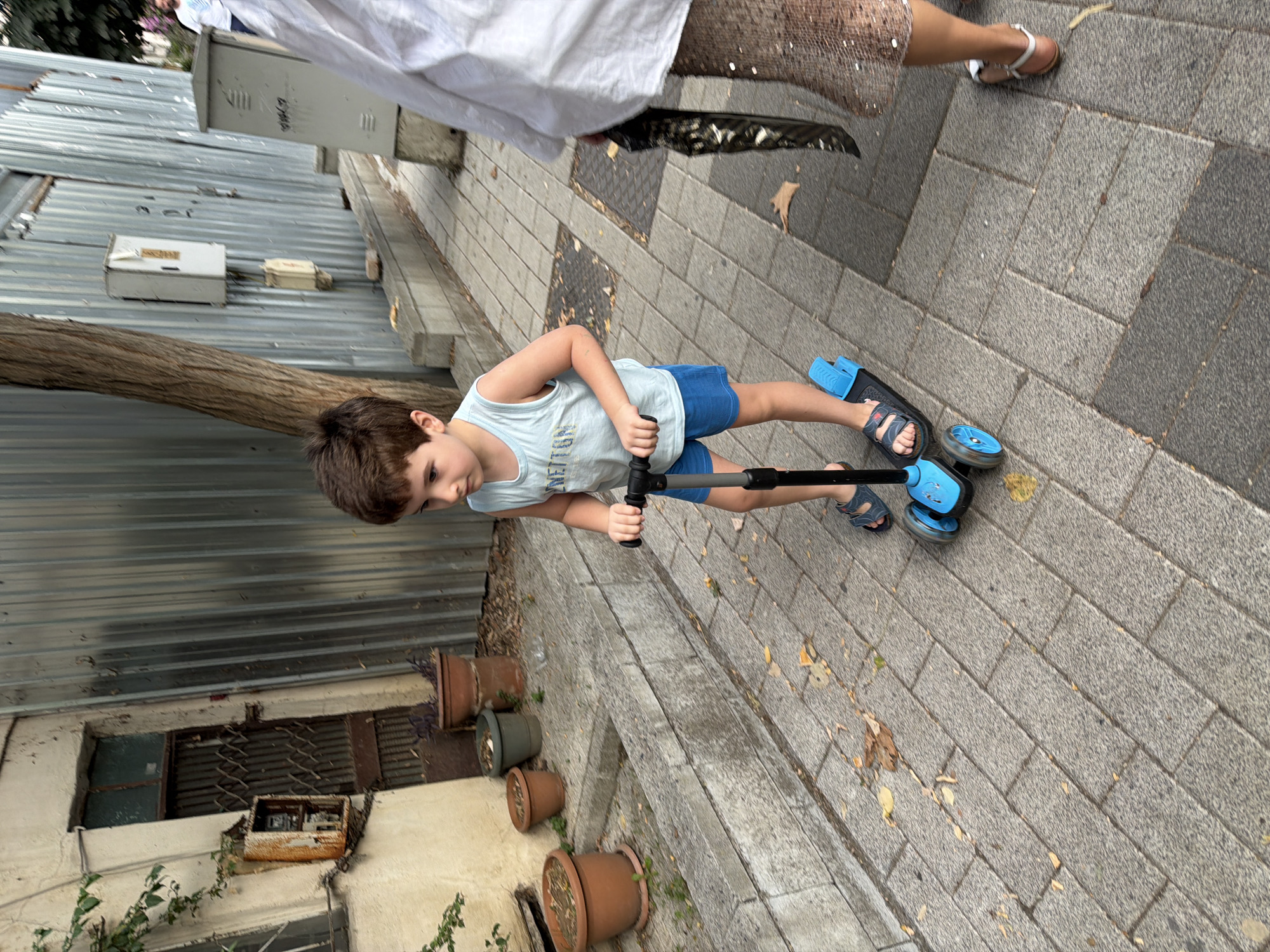 In Üsküdar, Turkey, a young boy with brown hair is riding a blue scooter on a sidewalk paved with gray cobblestones. He is wearing a light blue sleeveless shirt and blue shorts, with dark sandals on his feet. To the right of the boy, the lower half of a woman is visible. She has on a sparkly, sequined skirt and is holding a black bag. The background features a textured metal wall, a tree trunk, a building with an old door and a rusty electrical box, and several terracotta pots with plants. The scene appears to be outdoors in an urban setting.