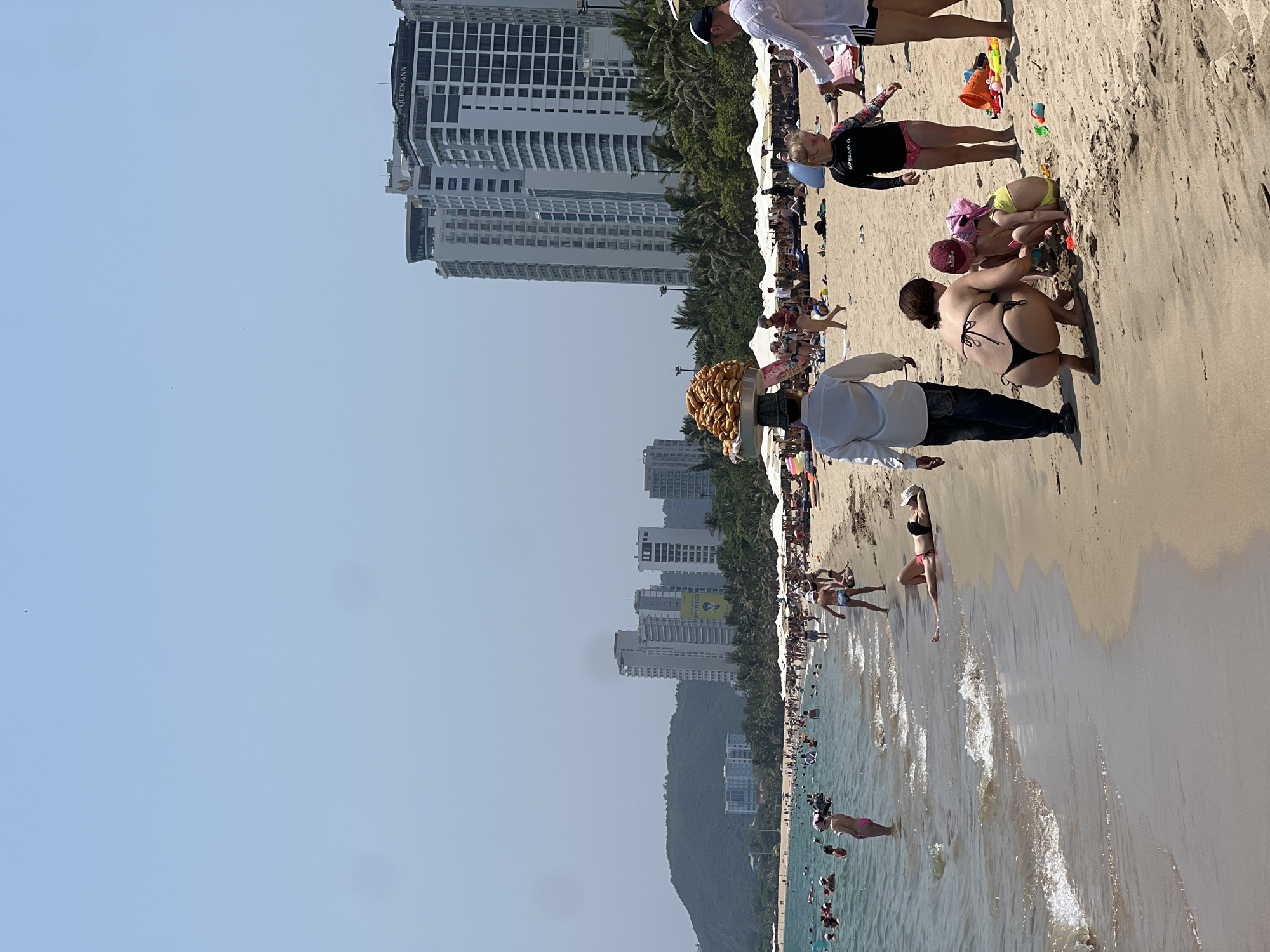 The image shows a crowded beach scene in Nha Trang, Vietnam. The beach is populated with numerous people, including families, individuals swimming, and beachgoers engaged in various activities. A vendor carrying a large tray filled with food approaches a group of people on the beach. The backdrop features high-rise buildings and lush green hills. The weather appears sunny and clear. The beach is lined with white umbrellas and towels placed on the sand. The sea is filled with people enjoying the water.