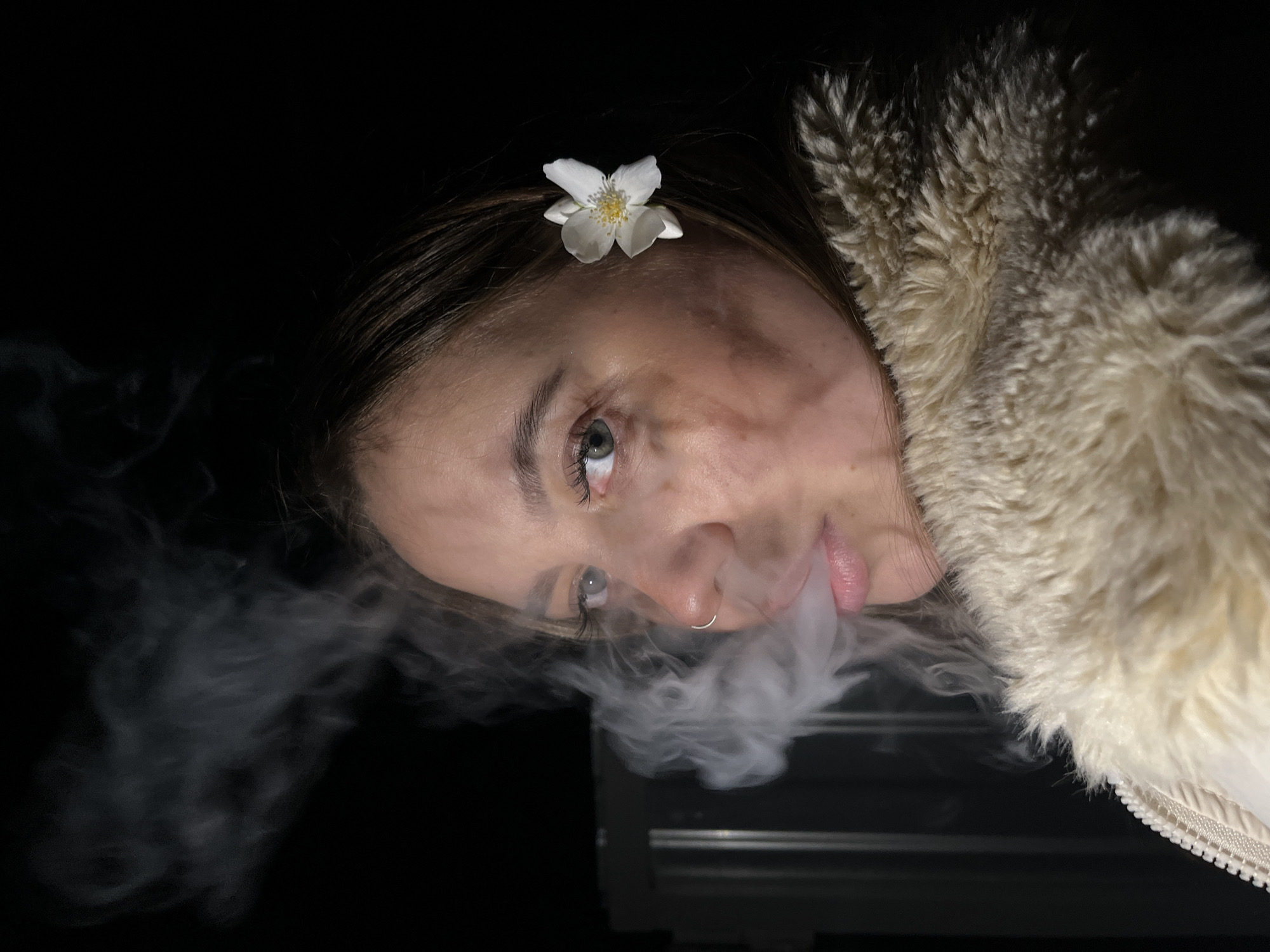 A close-up portrait shows a young woman against a dark, featureless background, suggesting nighttime or a dimly lit setting. The image is rotated approximately 90 degrees counter-clockwise, presenting her head tilted to the left. She has light skin, dark brown hair, and light-colored eyes, with long eyelashes and neatly groomed eyebrows. A small white flower with a yellow center is tucked into her hair above her forehead. A silver ring adorns her right nostril.
The primary activity depicted is the woman exhaling a cloud of dense white smoke from her mouth, which billows around her lower face before dissipating into the darkness. She is wearing a cream-colored jacket with a prominent, shaggy faux fur or fleece collar that frames the right side of her face. Her gaze is directed slightly upwards and to the left (relative to the image's current orientation), and her expression is neutral. The lighting is focused on her face and the smoke, creating a stark contrast with the dark background. Faint, indistinct horizontal lines are visible in the lower right corner, possibly part of an object or structure. No text is visible. The location context for this image is Novopokrovskaya, Russia.