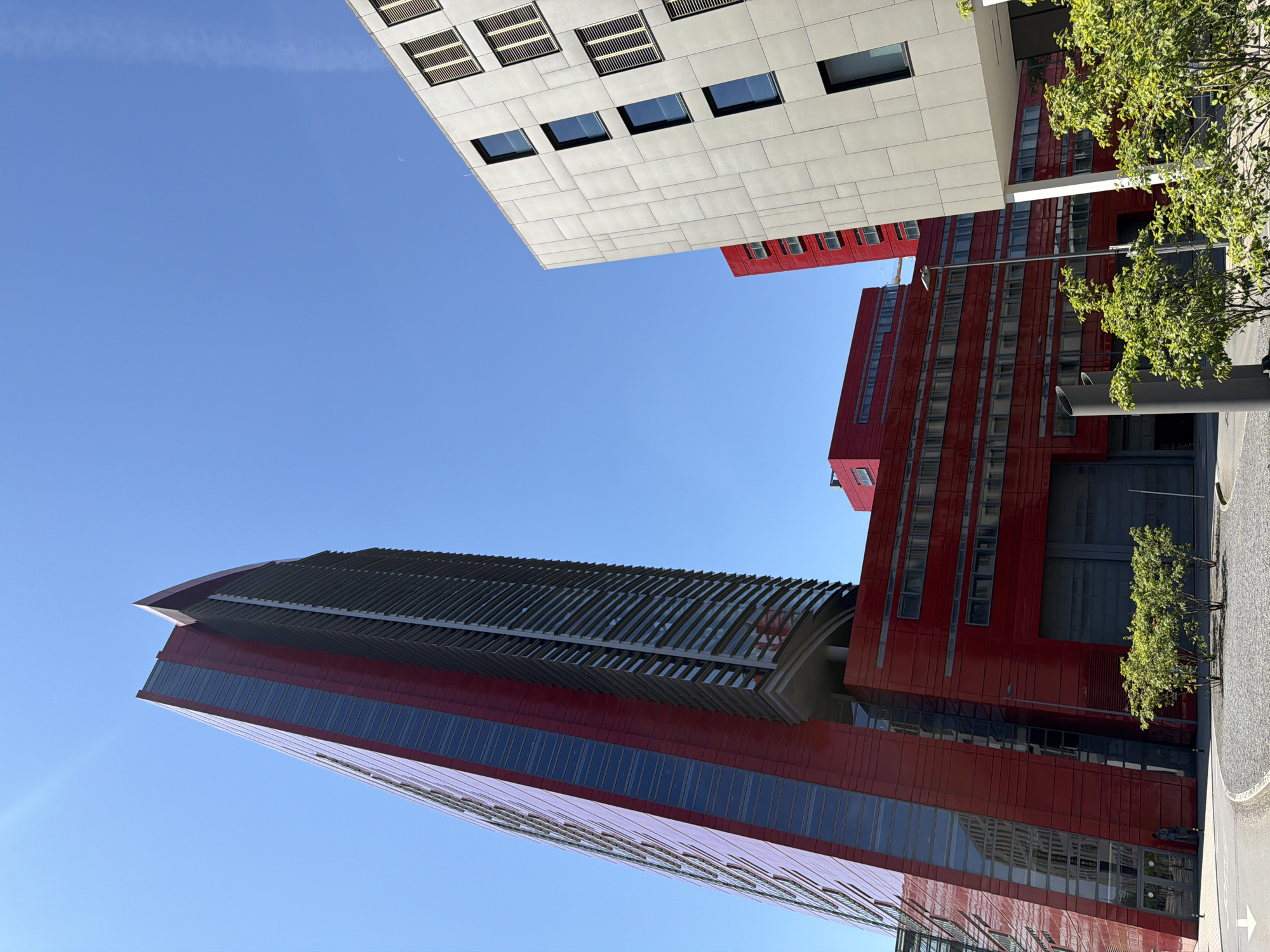 In Esch-sur-Alzette, Luxembourg, the image captures a modern architectural ensemble dominated by a towering, slender red skyscraper with a distinctive pointed top, its facade a blend of reflective red panels and dark vertical louvers. Adjacent to it stands a lighter-colored, multi-story building with a grid-like pattern of windows, some featuring horizontal blinds. In the foreground, a lower, elongated red structure with large, dark gray garage doors and rows of windows is visible. Small, leafy green trees are planted in a gravel-covered area near the base of the buildings. The scene is set against a clear, bright blue sky. The perspective is from a low angle, looking upwards, emphasizing the scale and geometry of the architecture.