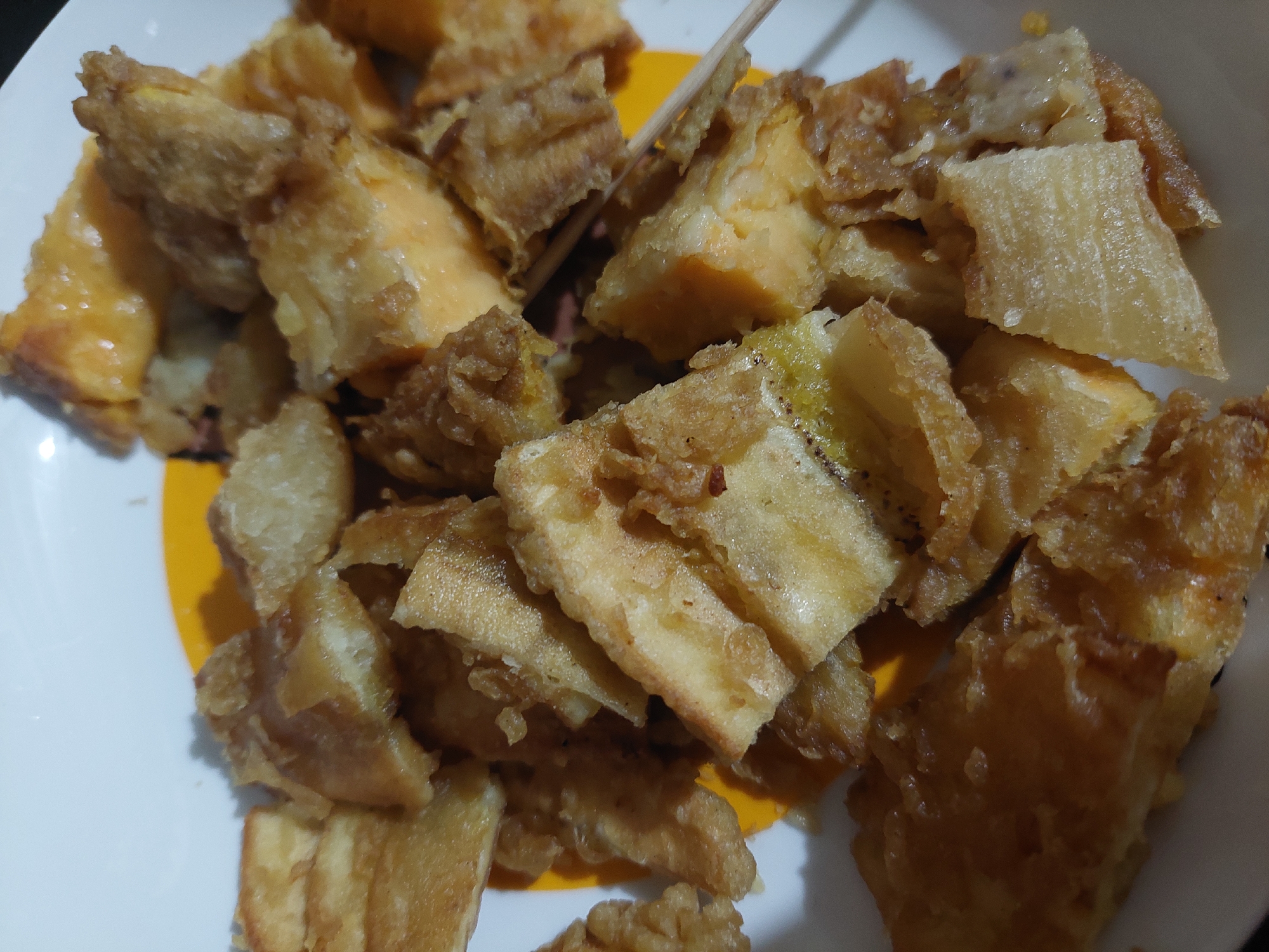 A close-up, overhead shot shows a plate of fried sweet potato pieces. The pieces are irregular in shape and size, with a golden-brown, crispy coating. Some pieces reveal the orange interior of the sweet potato. The food is arranged on a white plate with a yellow circular design. The overall lighting is dim, creating shadows on the food. The location is the city of Jelutung, Indonesia.