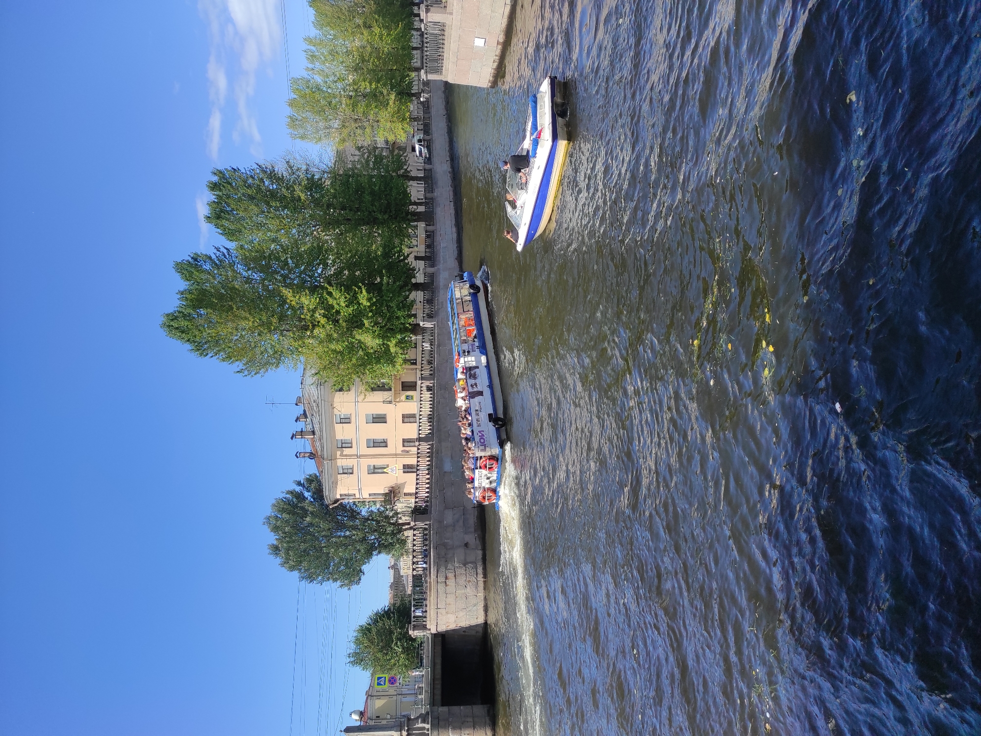 This is a medium shot, taken from a bridge overlooking a canal, of two boats navigating the water. The canal is bordered by stone embankments and a pedestrian walkway with railings. On the left embankment, a stone bridge with arches crosses the canal, and behind it, a yellow building with multiple windows and chimneys can be seen. Trees with green foliage line the canal banks, and the sky is a clear blue with a few wispy clouds.
The main subject is a large, white tour boat with a blue stripe along its side, filled with passengers who are standing and looking around. The boat is moving from left to right, leaving a wake in the dark, rippling water. The words "LION" and other illegible text are visible on the side of the boat. To the right of the tour boat, a smaller, white speedboat with a blue stripe is also moving from left to right. Two individuals are seated in the speedboat; one is operating the boat from the helm, and another is a passenger.
The overall impression is a sunny day in Saint Petersburg, Russia, with the canal being a popular spot for boat tours. The scene captures a moment of leisure and sightseeing.