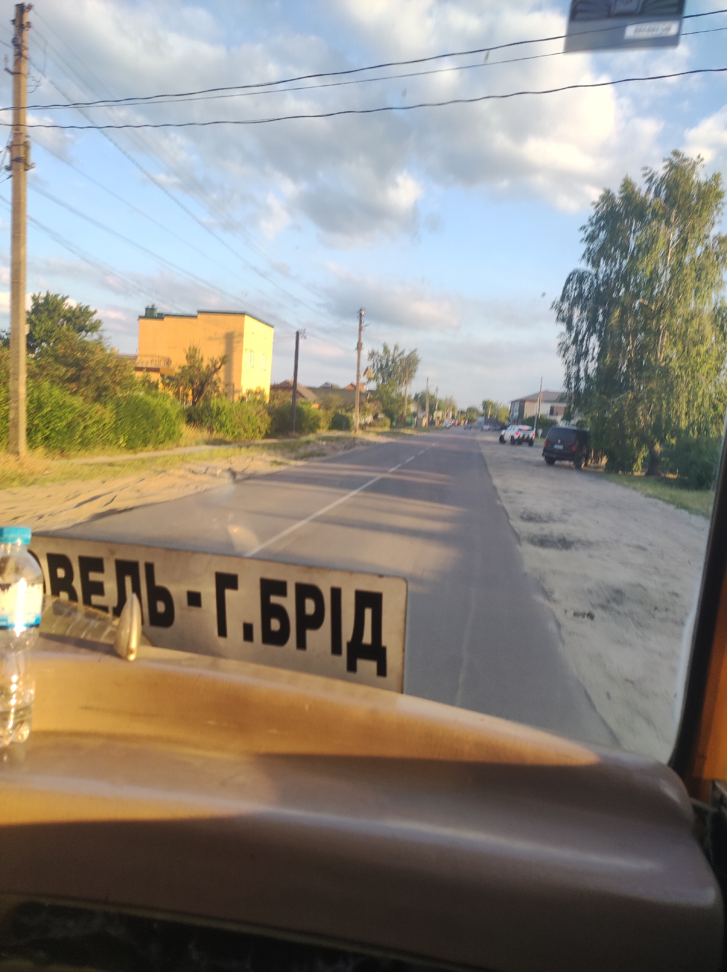 The image shows a road with a yellow building and trees on the side, as well as utility poles and power lines overhead. In the foreground, there is a yellow surface with a sign that reads "ВЕЛЬ - Г.БРІД". A plastic bottle of water is also visible on the yellow surface. In the distance, there are cars on the road and more buildings. The sky is cloudy with patches of blue. The scene appears to be in a rural or suburban area during the daytime.