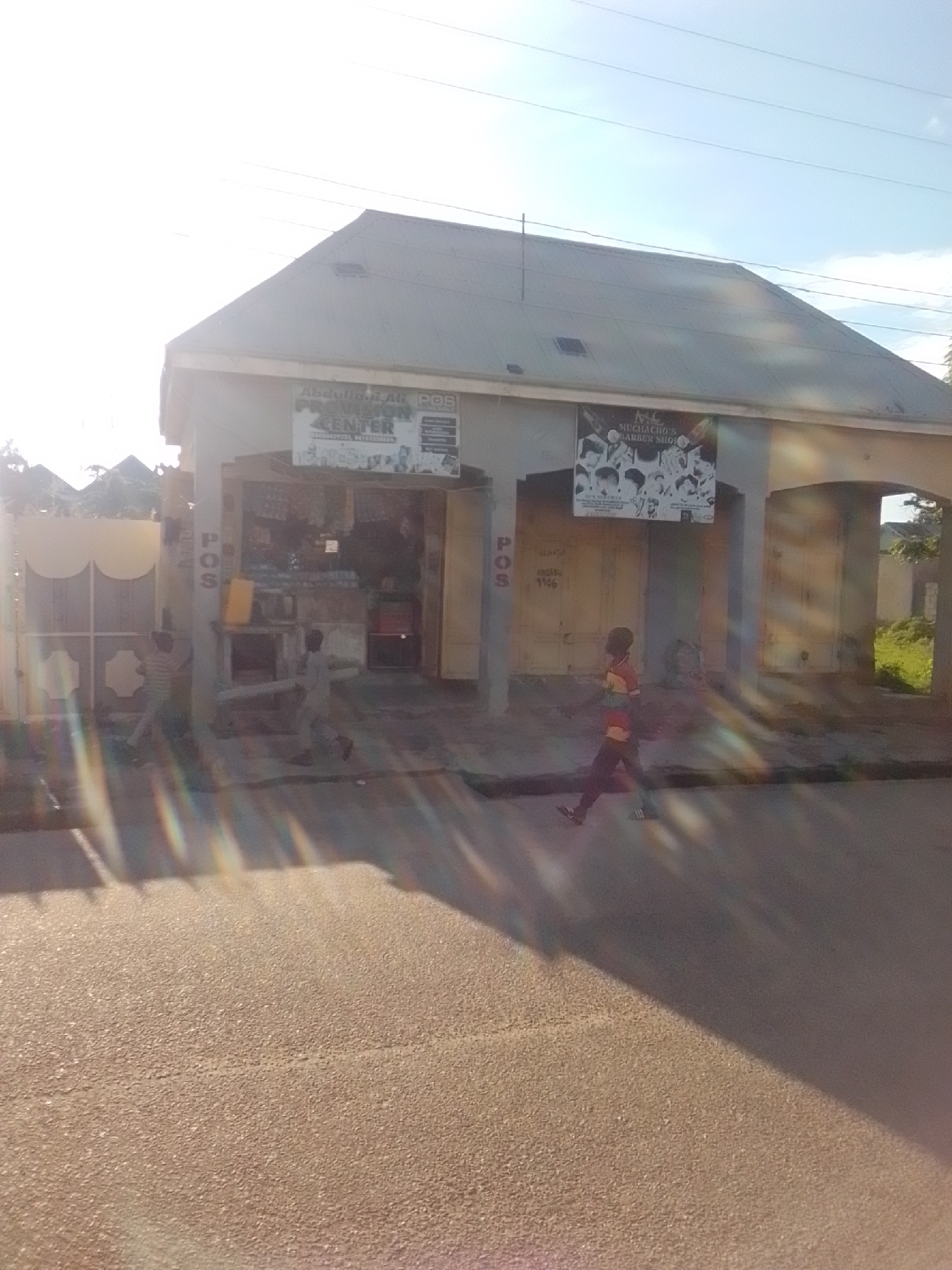 The image captures a street scene in Gombe, Nigeria, likely near a commercial area. In the foreground, a paved road is visible, with sunlight casting long shadows and lens flares across the surface. The middle ground features a single-story building with a gray, sloped roof. The building appears to be a commercial establishment, possibly a shop or a collection of small businesses.
Two signs are prominently displayed on the front of the building. One sign, in green and white, reads "ABDULLAHI AB POS PROVISION CENTER" and lists contact numbers. Another sign, with a black background and white lettering, advertises "MC MUCHAHO'S BARBER SHOP" and displays images of various hairstyles. Below these signs, the storefront appears to be open, with shelves and items stocked inside, suggesting a provision store or similar retail outlet. There are also vertically oriented "POS" signs on pillars of the building.
In front of the building, two young boys are captured in motion. One boy, wearing a striped shirt and dark pants, is walking towards the left of the frame. Another boy, wearing a red and white shirt and dark pants, is running towards the right of the frame. A yellow container is visible near the entrance of the provision center.
The background shows more of the building, including arched openings, and hints of trees and vegetation to the right. The sky is partly cloudy with visible power lines crisscrossing overhead. The overall impression is a snapshot of daily life in a bustling commercial part of Gombe.