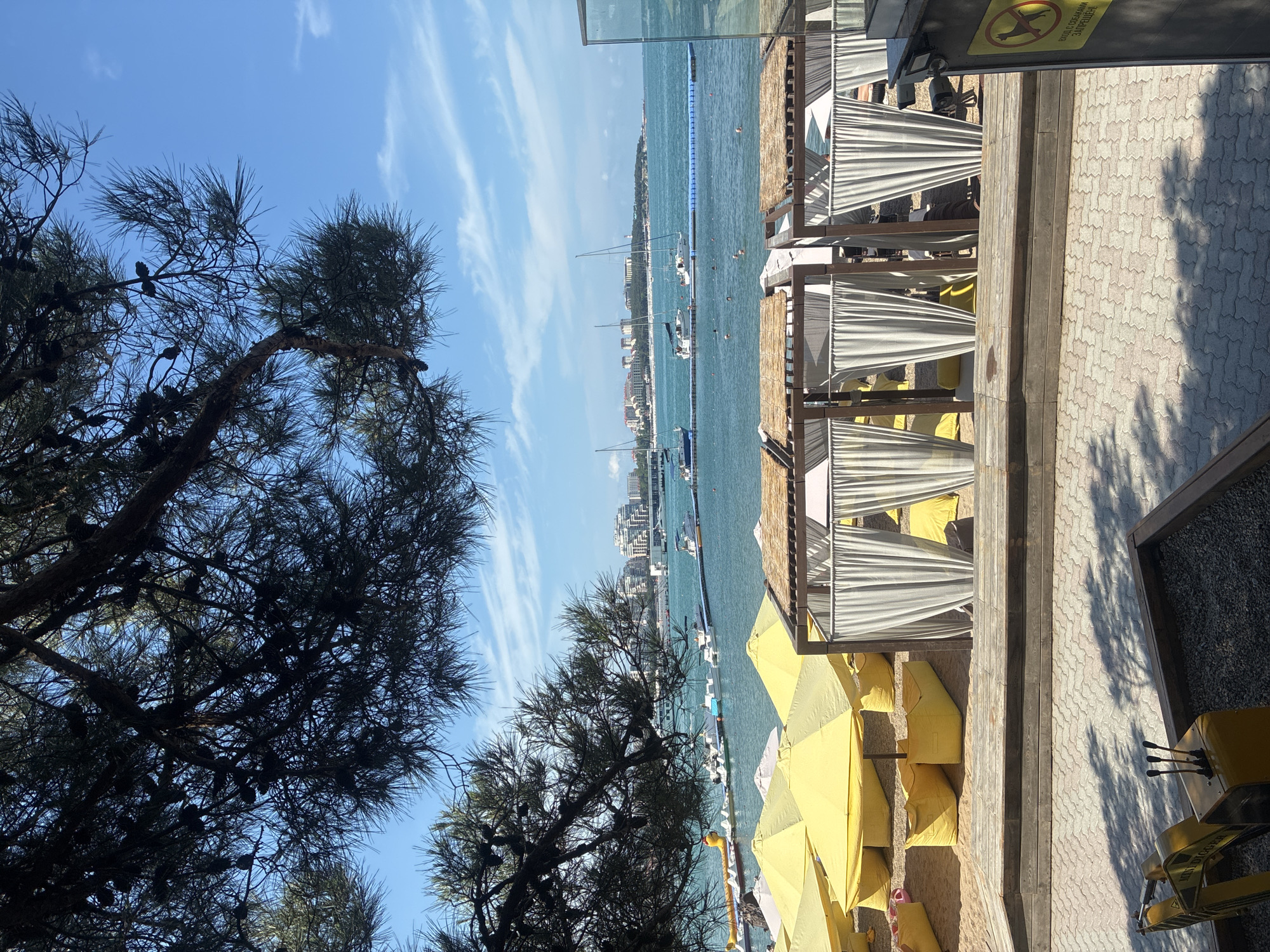 The image depicts a sunny day at a beach resort, likely located in Gelendzhik, Russia, based on contextual clues. In the foreground, a yellow toy excavator sits in a sandbox. Behind it, a paved walkway is shaded by the branches of a large pine tree. Further back, a beach area is set up with numerous yellow umbrellas and beanbag chairs. Several covered cabanas with white curtains are also visible. Beyond the beach, the turquoise water of the sea stretches to the horizon, dotted with several sailboats and a long blue floating barrier. In the distance, a coastal city skyline with buildings and a lighthouse is visible against a clear blue sky with scattered clouds. Some people can be seen swimming in the sea.