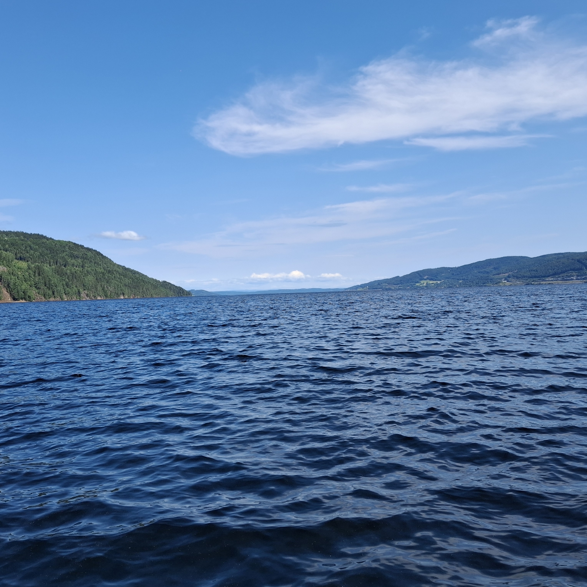The image depicts a panoramic view of a large body of water, likely a lake or a fjord, under a clear blue sky with scattered white clouds. In the foreground, the dark blue water exhibits gentle ripples and waves, reflecting the sky. On the left side of the frame, a densely forested hillside slopes down towards the water. The trees are predominantly evergreen, creating a lush green expanse. In the distance, the landscape is characterized by rolling hills and mountains, some of which are also covered in vegetation. The far horizon shows faint outlines of more distant landmasses. The overall scene conveys a sense of natural beauty and tranquility, suggesting an outdoor recreational setting. The location is identified as Minnesund, Norway, which is known for its scenic waterways and surrounding natural landscapes. There are no people, animals, or man-made structures immediately visible in the frame, focusing the attention on the natural environment.