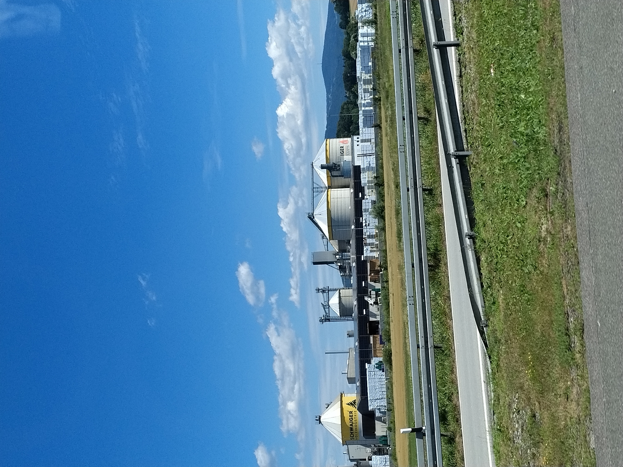 The image shows an industrial facility under a bright blue sky with scattered white clouds. The facility appears to be a biomass pellet plant, as suggested by the signage on one of the silos which reads "SCHWAIGER ROLL INDUSTRIE" and another with "GER PELLETS". There are large, conical silos, storage buildings, and stacks of bundled material, possibly wood pellets or lumber, outside. The plant is situated in a rural area, with a field of dry grass in the foreground and a wooded hillside in the background. A highway guardrail runs across the middle ground, separating the viewer from the industrial site. The image appears to have been taken from a moving vehicle on the highway. The location is identified as Hengersberg, Germany.