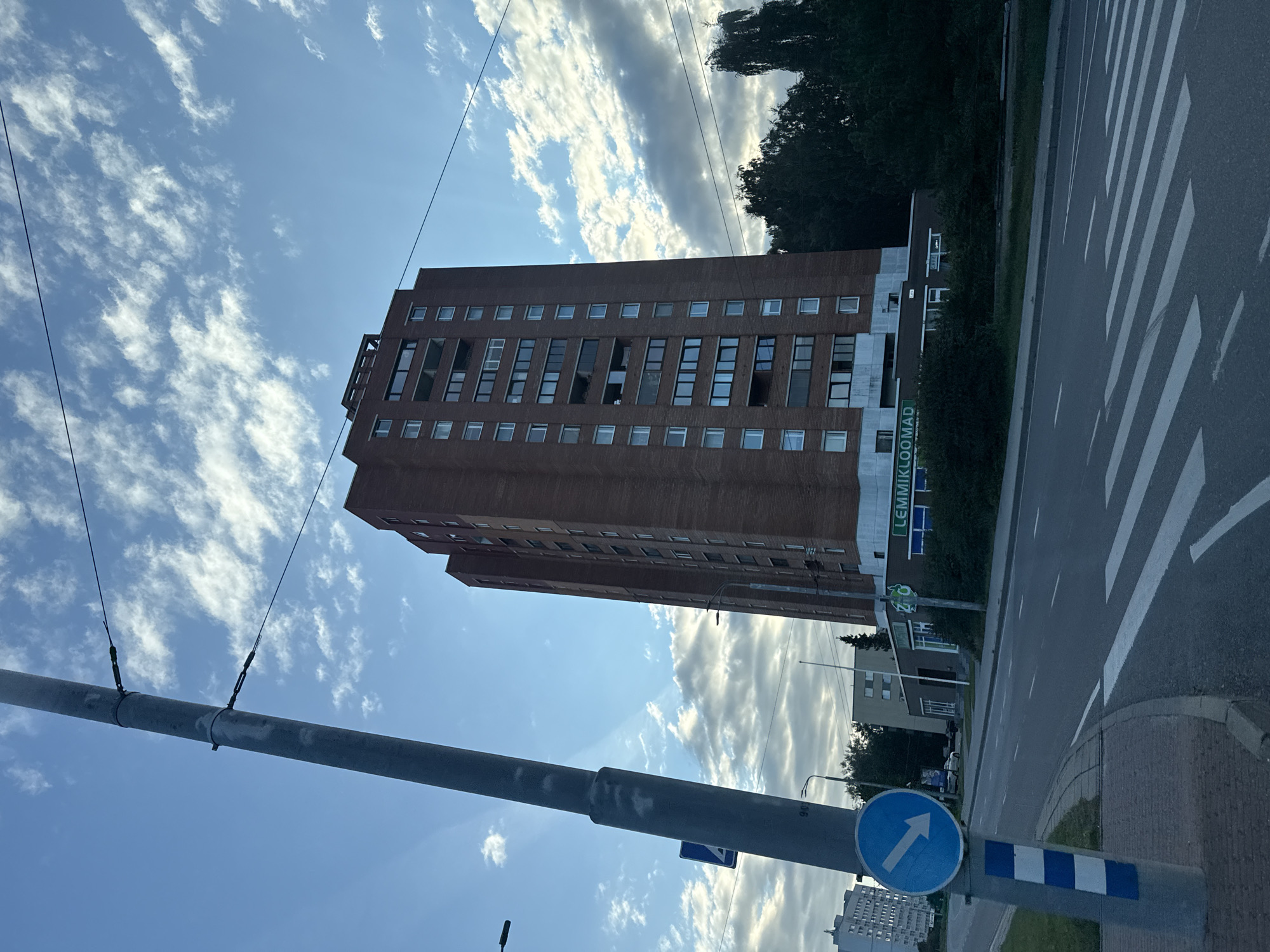 This is a daytime, eye-level shot of a tall, brick apartment building in Tallinn, Estonia, viewed from a street. The building is a dominant feature, characterized by its numerous windows and balconies. In the foreground, a gray utility pole with blue and white stripes at its base stands to the left. A blue circular traffic sign with a white arrow pointing right is attached to the pole. A crosswalk with white stripes is visible on the asphalt road in front of the building. To the right of the building, a dense line of dark green trees can be seen. The sky above is a mix of blue with scattered white clouds. A low-rise commercial building with the sign "LEMMIKLOOMAD" (meaning "pets" in Estonian) is situated at the base of the apartment building. Other buildings and streetlights are present in the background, suggesting an urban environment.