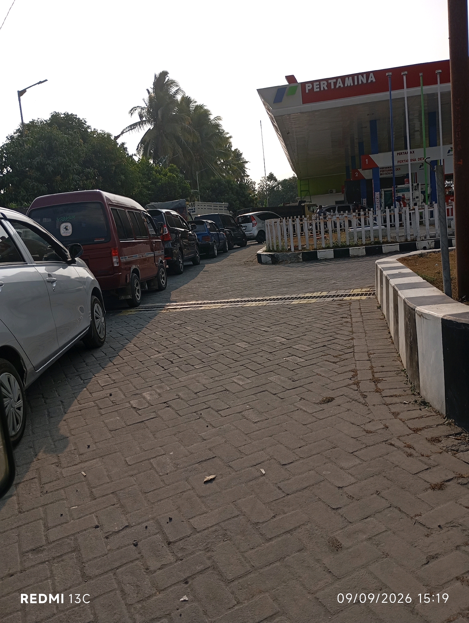 The image shows a line of vehicles at a Pertamina gas station in Rowotamtu, Indonesia, under a bright, overcast sky. In the foreground, a silver car is parked on a brick-paved surface. Behind it, a red van and several other cars are lined up, seemingly waiting to refuel. The Pertamina station's distinctive red and white signage is visible in the background, along with multiple gas pumps. The surrounding environment includes lush green trees and palm trees, suggesting a tropical or subtropical climate. The ground is paved with interlocking bricks, and a black and white striped barrier runs along the edge of the paved area. The timestamp "09/09/2026 15:19" is overlaid on the bottom right of the image, and "REDMI 13C" is on the bottom left.