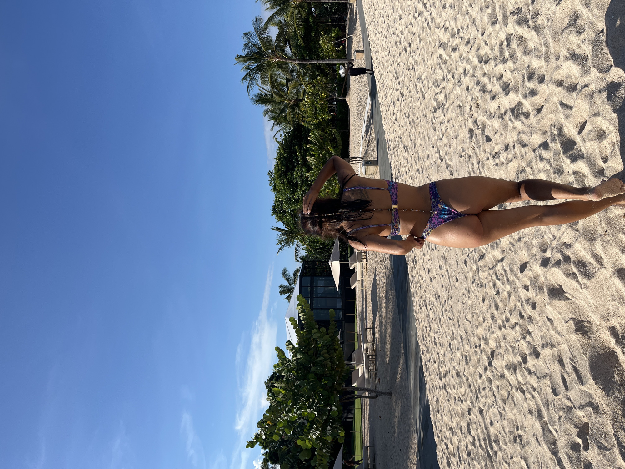 The image displays a woman from a rear perspective, walking or posing on a sun-drenched sandy beach in Kuta Selatan, Indonesia. She is wearing a vibrant purple and blue patterned bikini with intricate strap designs and beaded embellishments. Her dark hair appears to be styled in braids or dreadlocks, some visible over her shoulder, with her right hand near the back of her head, perhaps adjusting her hair or bikini.
The scene is a tropical coastal environment characterized by extensive, light-colored sand marked with footprints. Lush green tropical vegetation, including towering palm trees and dense bushes, lines the left side of the frame, separating the beach from background structures. Beyond the foliage, modern-style resort buildings with large windows and a white tent-like structure are visible. Another person, appearing as a small dark silhouette, can be seen further down the beach near the resort facilities.
The sky is a clear, bright blue with minimal, wispy clouds, indicating excellent weather conditions. The strong sunlight casts long shadows across the sand from the woman and unseen elements, suggesting it is either mid-morning or early afternoon. The overall atmosphere conveys relaxation and a luxurious beach destination. There is no visible text in the media file.
