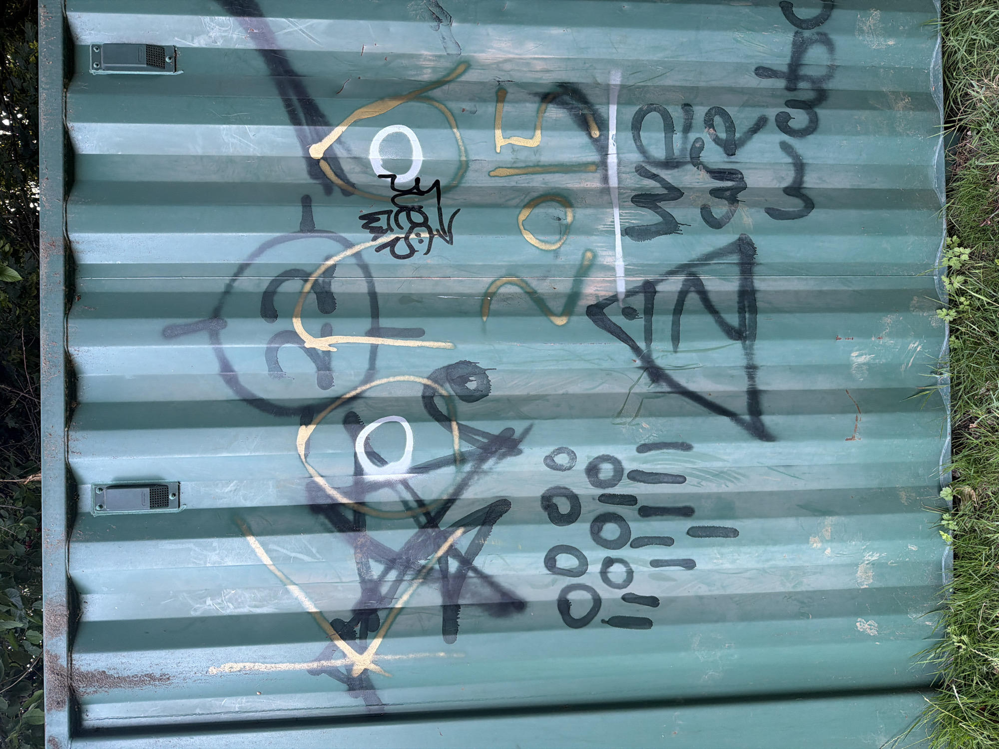 This image displays a section of a vertical, corrugated metal structure, painted a dark teal or green, situated in an outdoor environment. The surface exhibits significant weathering, including streaks of dirt, grime accumulation, and minor areas of rust, particularly along its upper edge.
Two rectangular, dark grey plastic devices, each secured by two visible screws, are mounted on the upper portion of the corrugated panel. These devices appear to be vents or small enclosures; the one on the left is partially obscured by accumulated dirt.
The metal surface is extensively covered with multiple layers of graffiti. Key elements include:
* An abstract design towards the upper-left, comprising black spray paint lines forming an 'X' and other shapes, intertwined with a gold circular outline. A small white ring is visible within this complex.
* In the upper-right section, a stylized "eye" or "face" motif is present, featuring a gold outline and a prominent white circle for the eye. Above this, a black spray-painted tag, resembling "bang" with a crown symbol, is visible.
* Below the upper-left abstract graffiti, an arrangement of multiple black spray-painted circles of varying sizes is present, below which several vertical black lines, indicative of paint drips, are visible.
* The numbers "2015" are spray-painted in gold in the middle-right area of the panel.
* A thick, horizontal white spray-painted line runs across the panel beneath the "2015" numbers.
* Below the white line, in the bottom-right, the partial phrase "we are watc" (suggesting "we are watching") is rendered in black spray paint, also showing drips.
* Towards the bottom-left, a large black spray-painted triangle containing a stylized 'Z' or similar symbol is depicted, crossing the horizontal white line.
The top of the image reveals dark green foliage, consistent with trees or bushes, against a lighter background. At the base of the metal structure, green grass and some sparse weeds are visible, meeting the ground level. The diverse styles and colors of graffiti suggest applications over different periods. The scene is consistent with an urban or semi-urban outdoor setting, as found in a city like Maidstone, United Kingdom.