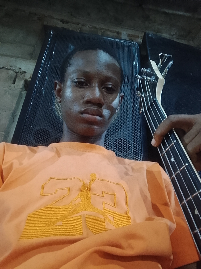 A young man in an orange t-shirt featuring a yellow "23" design takes a selfie. He's holding the neck of a bass guitar, strings and tuning pegs are visible. A large black speaker with a mesh front sits behind him to his left, and another speaker is partially visible on his right. The lighting is dim, creating shadows on his face and the surrounding objects.