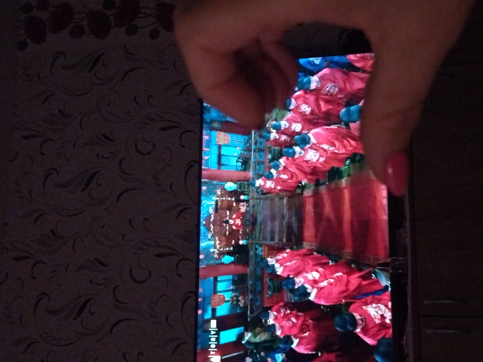 The image shows a vertical view of a smartphone displaying a scene of a group of people wearing red shirts and blue caps, presumably in a public area or event. The background suggests architecture with blue and red elements. A hand with pink nail polish is partially covering the screen. The location is possibly Stara Zagora, Bulgaria, based on the user's prompt. The environment includes a patterned wall to the left and a dark wooden surface to the right.
