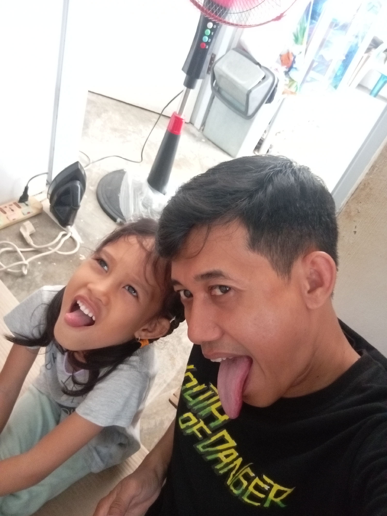 A selfie taken by a man and a young girl shows them sticking out their tongues. The man, on the right, wears a black t-shirt with green lettering that appears to say "SOUTH OF DANGER". The girl, on the left, is wearing a gray t-shirt and light blue pants. Her tongue is also extended. In the background, a black and red standing fan is visible, along with an iron and a power strip on the floor. The environment appears to be a room with tiled flooring and white walls, possibly a residential area in Batam, Indonesia.