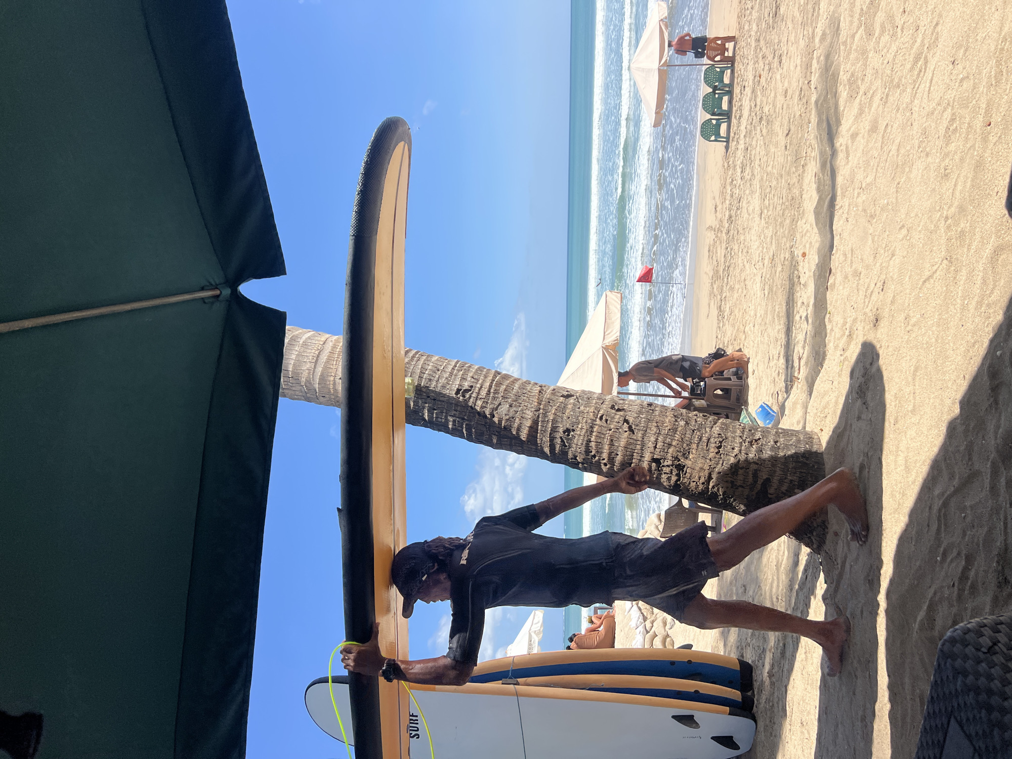 A man wearing a black wetsuit and shorts carries a surfboard overhead while walking on a sandy beach in Kuta, Indonesia. He is walking past a palm tree and several other surfboards. In the background, the ocean stretches out to the horizon under a clear blue sky. Beach umbrellas and chairs are visible, along with other people enjoying the beach. The scene is sunlit and appears to be a warm, clear day.