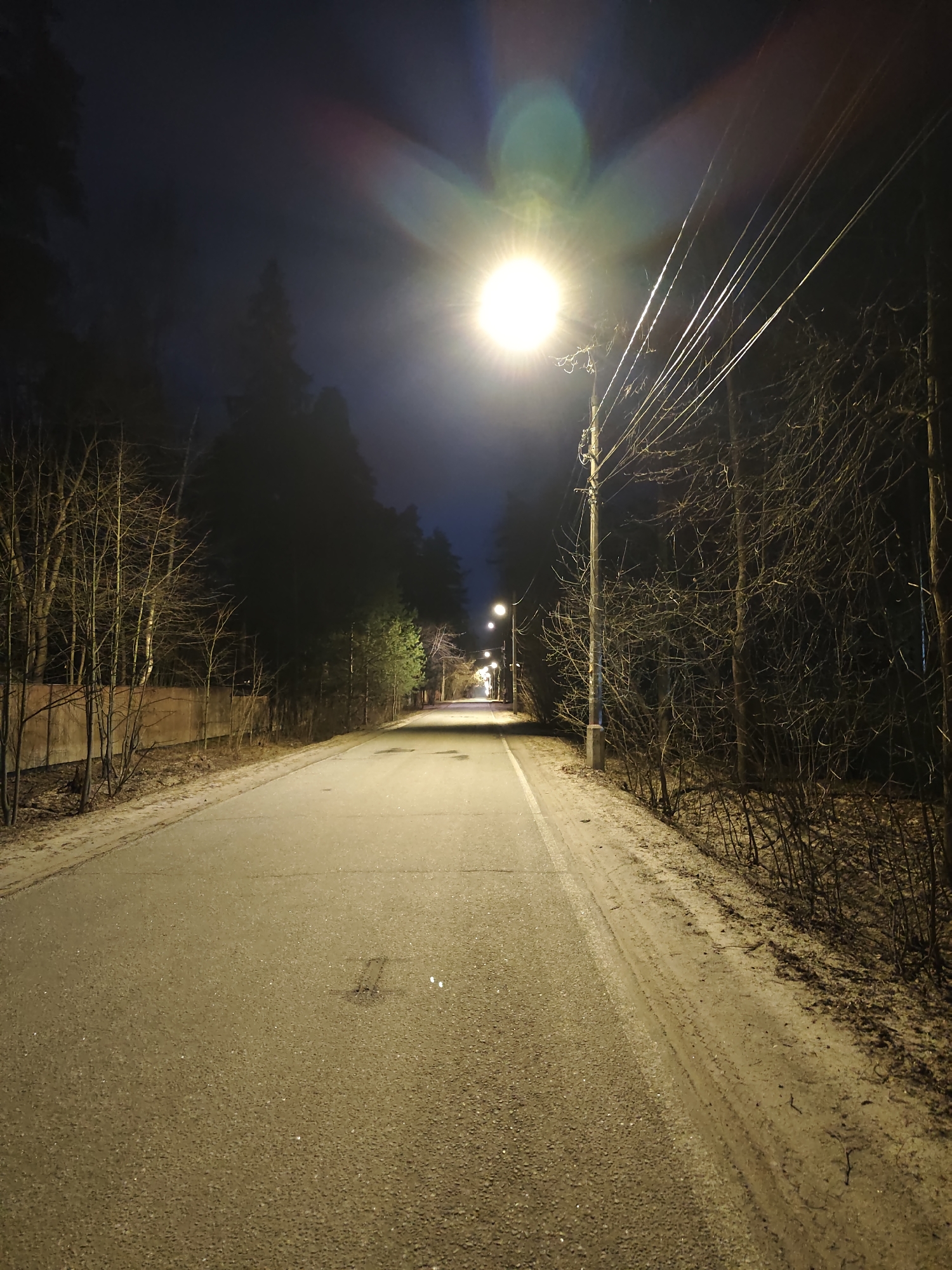 The image depicts a paved road in what appears to be a residential area. It is night, and illumination is provided by streetlights. The road leads into the distance. On either side of the road are trees and vegetation, mostly bare, suggesting a late fall or winter season. There are power lines visible, strung from a utility pole. Based on the presence of what looks like a concrete fence or wall along the left side of the road, there are residential properties bordering the road. There is a faint sign of what could be tire tracks or a discoloration on the road surface. The context provided suggests the location is the city of Kratovo, Russia, however, this cannot be verified from the image alone. No people, subjects, actions or interactions are visible.