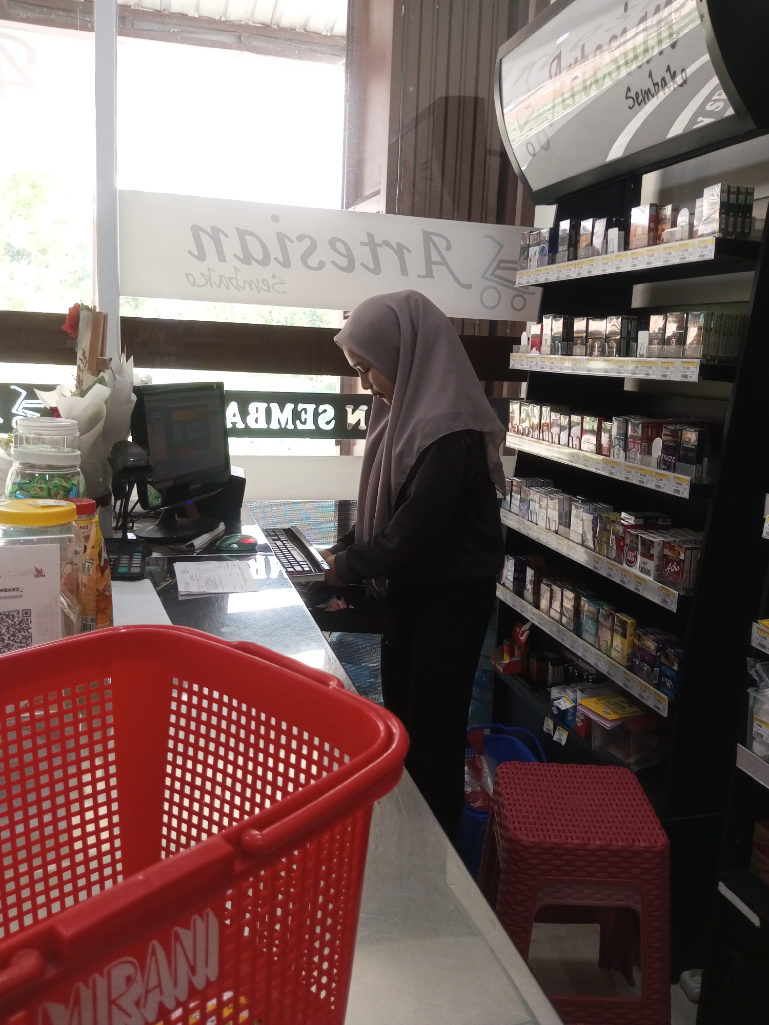 The image captures a scene inside a store, likely a convenience store or minimarket, in Andongsili, Indonesia. In the foreground, a bright red plastic shopping basket is prominently displayed. To the right, a dark red woven plastic stool is visible. The midground features a counter area with a computer monitor, a barcode scanner, a calculator, and various items including jars, bottles, and a stack of receipts. A person wearing a light purple hijab and dark clothing is positioned behind the counter, interacting with the computer and keyboard, possibly as a cashier. In the background, shelves are stocked with numerous cigarette packs, indicating a wide selection available for purchase. Above the shelves, a large sign with stylized text is displayed. The overall environment suggests a commercial setting designed for retail transactions.