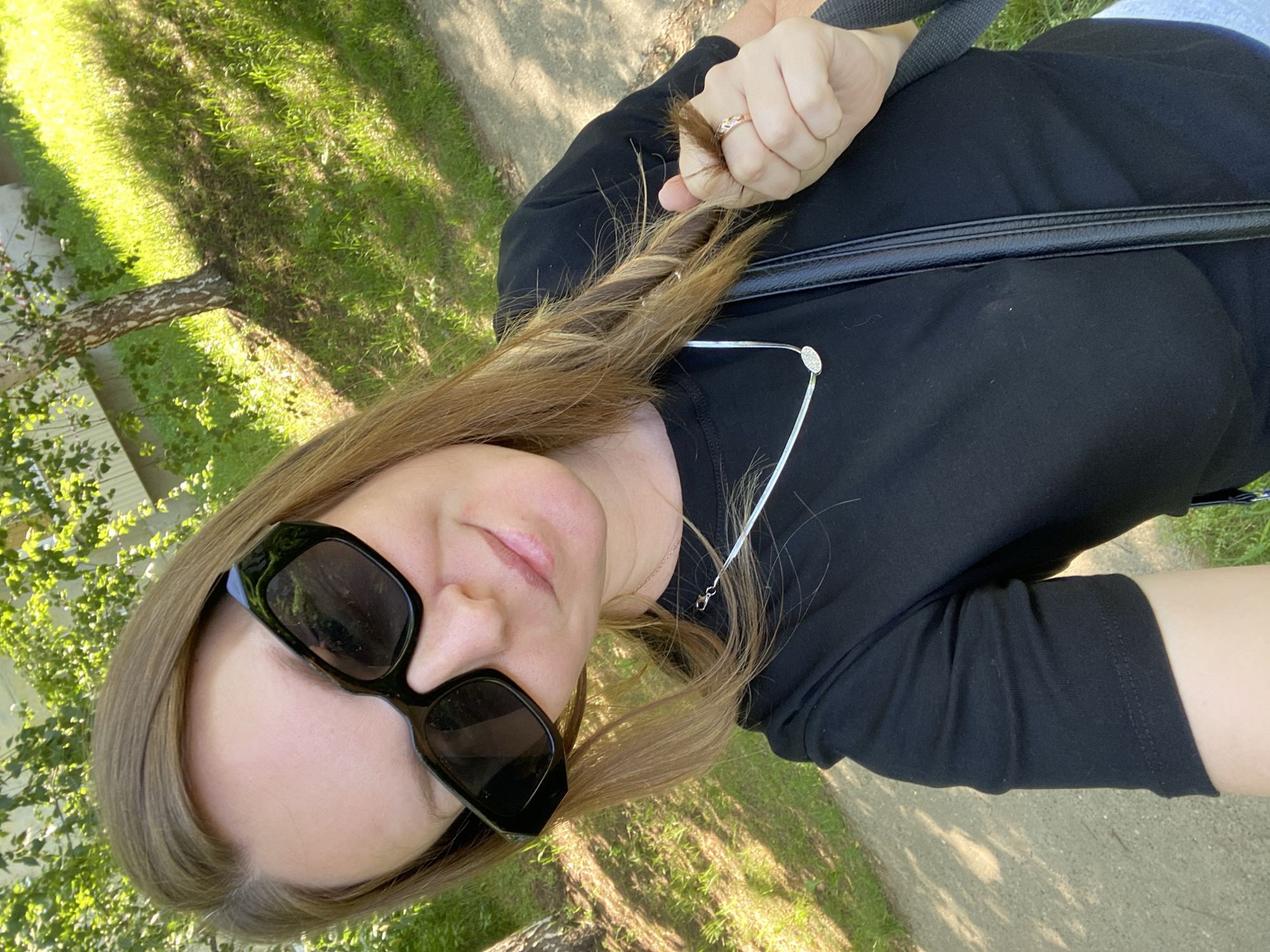 The image shows a selfie of a woman wearing black sunglasses, a black t-shirt, and a silver necklace. She is holding a strand of her brown hair with her right hand, on which she is wearing a ring. She is standing outdoors in what appears to be a park or green space with trees and grass. The ground is a paved path. The lighting suggests it is daytime, likely with sunlight filtering through the trees. There is no indication that this location is in Noviy, Russia, or any other specific city.