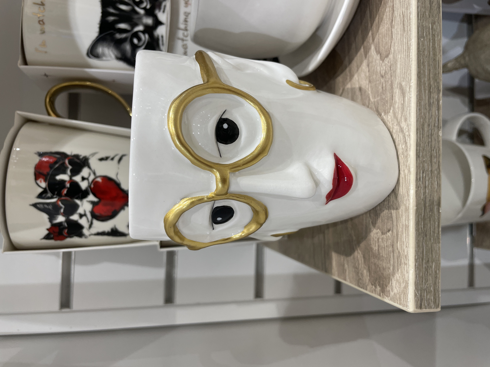This media file shows a close-up, rotated view of three ceramic mugs stacked on a shelf. The foreground mug is white with a stylized face, featuring large gold-rimmed glasses, black eyes, and red lips. The mug also has a gold handle. Behind it, another mug is visible, decorated with black and red hearts and a black and white illustrated cat wearing a red bow. The third mug, mostly obscured, is white and appears to have a gold handle. The mugs are displayed on a shelf with a wood-grain finish. The background is a plain white surface, likely a store display or wall. The lighting suggests an indoor setting, possibly a retail environment. There is no visible text.