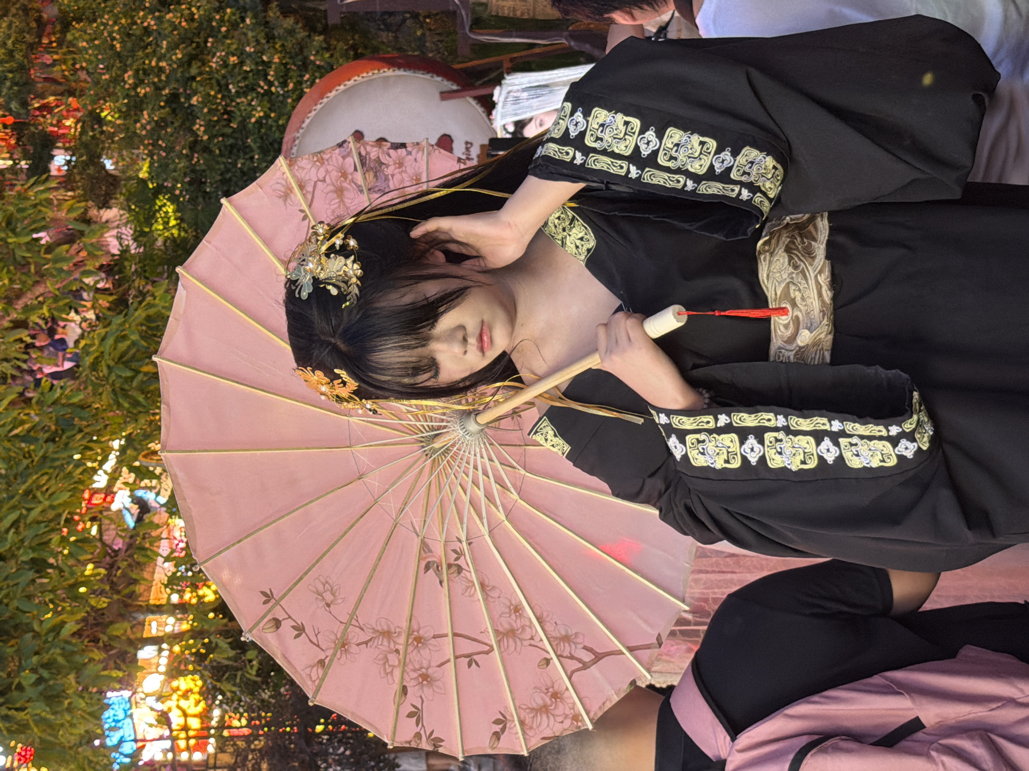 The image features a young woman in traditional Chinese attire, possibly a hanfu, holding a pink parasol. She has dark hair adorned with gold and green accessories and is looking down with a gentle expression. The parasol is open and decorated with a floral pattern. The woman is wearing a black robe with intricate gold embroidery on the sleeves and chest area.
The scene appears to be outdoors at dusk or night, given the presence of blurred colorful lights in the background, suggesting a festive or illuminated environment like a market or a celebration. The background also shows greenery and what looks like a large drum. The overall mood is serene and elegant, possibly capturing a moment during a cultural event or a photoshoot. The context provided indicates the location is Yuzhong District, China.