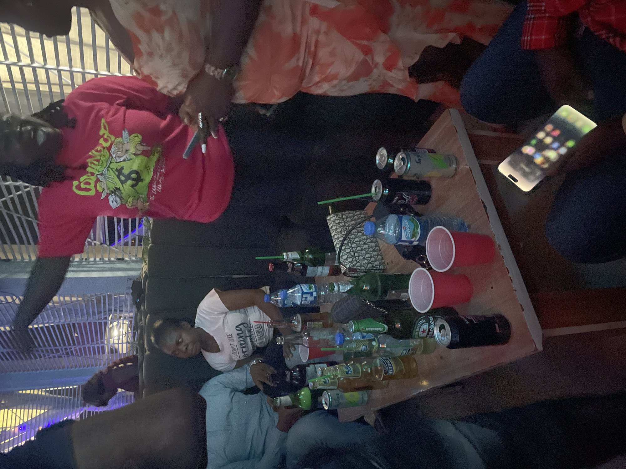 The scene depicts a group of individuals gathered around a table covered with numerous bottles and cans of beverages, including beer, water, and soft drinks. Red plastic cups are also present on the table. Several people are partially visible, their clothing indicating a casual setting. One person is holding a lit-up smartphone. The location, city of Jos, Nigeria, is not discernible from the visual information provided.