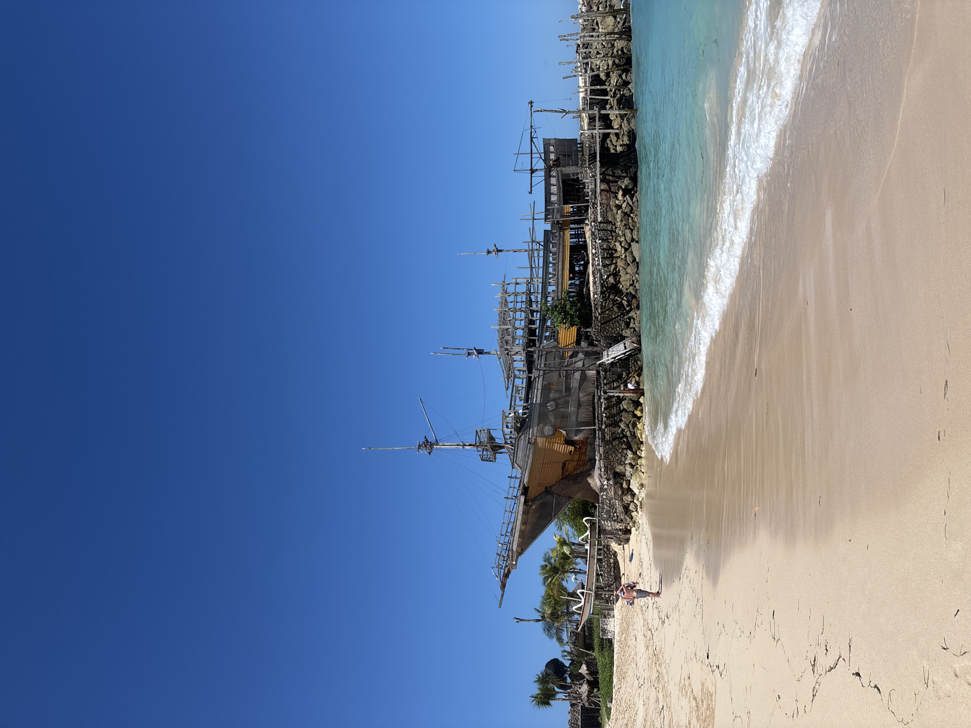 This image depicts a coastal scene in Kuta Selatan, Indonesia. A replica pirate ship is the central subject, positioned along a rocky coastline meeting a sandy beach. The sky is a clear, vibrant blue. On the beach, individuals are present, though their activities are not distinctly discernible. The water is a light turquoise color with visible wave action. Palm trees and some vegetation are scattered along the coastline near the ship. The artificial structure next to the beach could be the Pirate’s Bay restaurant.