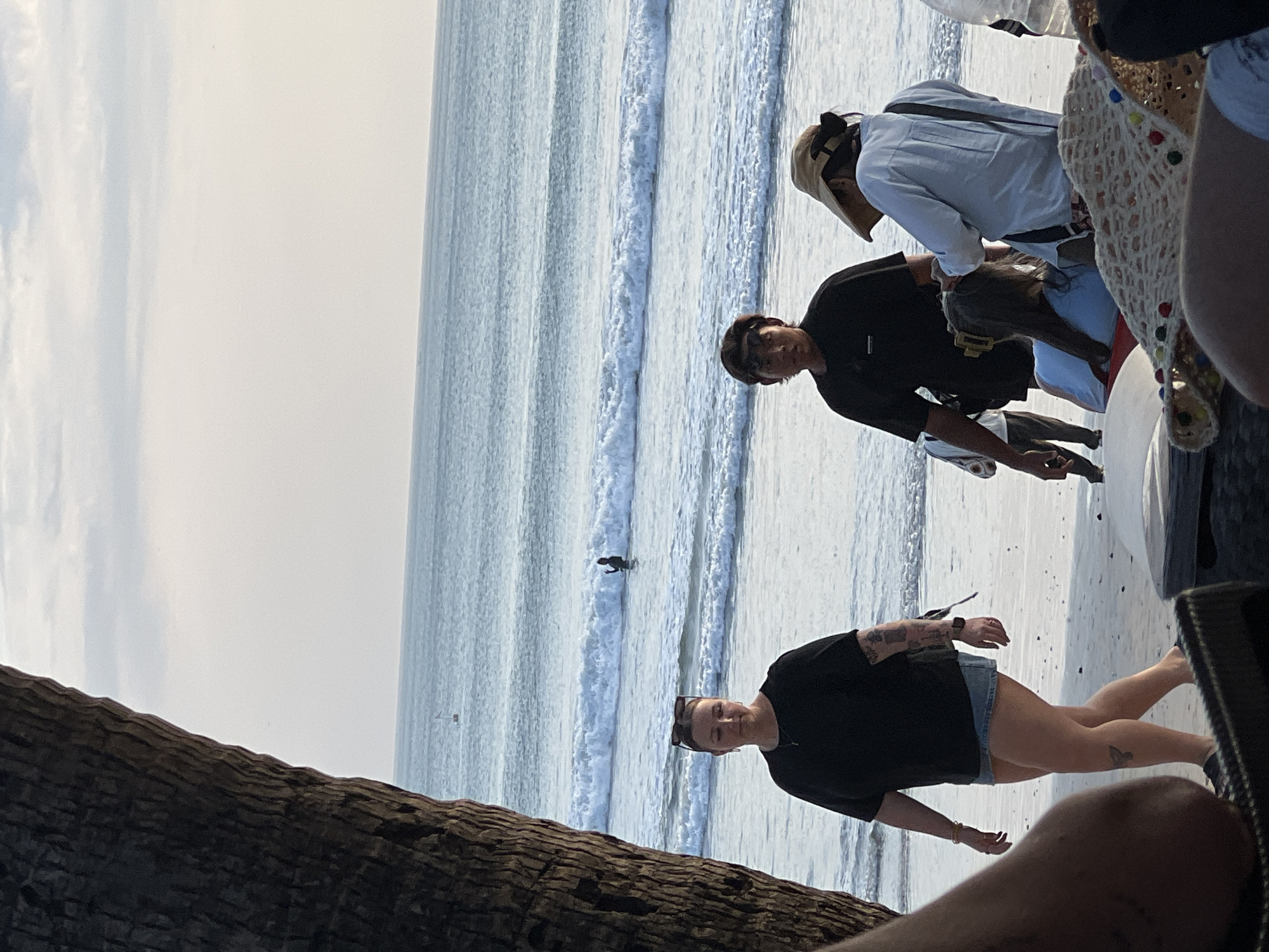 At a Kuta, Indonesia beach, several individuals are present. In the foreground, a woman with sunglasses on her head and tattoos on her arm walks towards the right, wearing a black t-shirt and denim shorts. To her right, a man wearing a black t-shirt stands facing the camera. Further right, a person wearing a light blue shirt and a wide-brimmed hat is seated, with another person in a light blue shirt facing away from the camera. In the background, a surfer is visible in the ocean, with gentle waves breaking on the shore. A large tree trunk partially obscures the left side of the image. The sky is overcast, and the lighting suggests it is late afternoon or early evening.