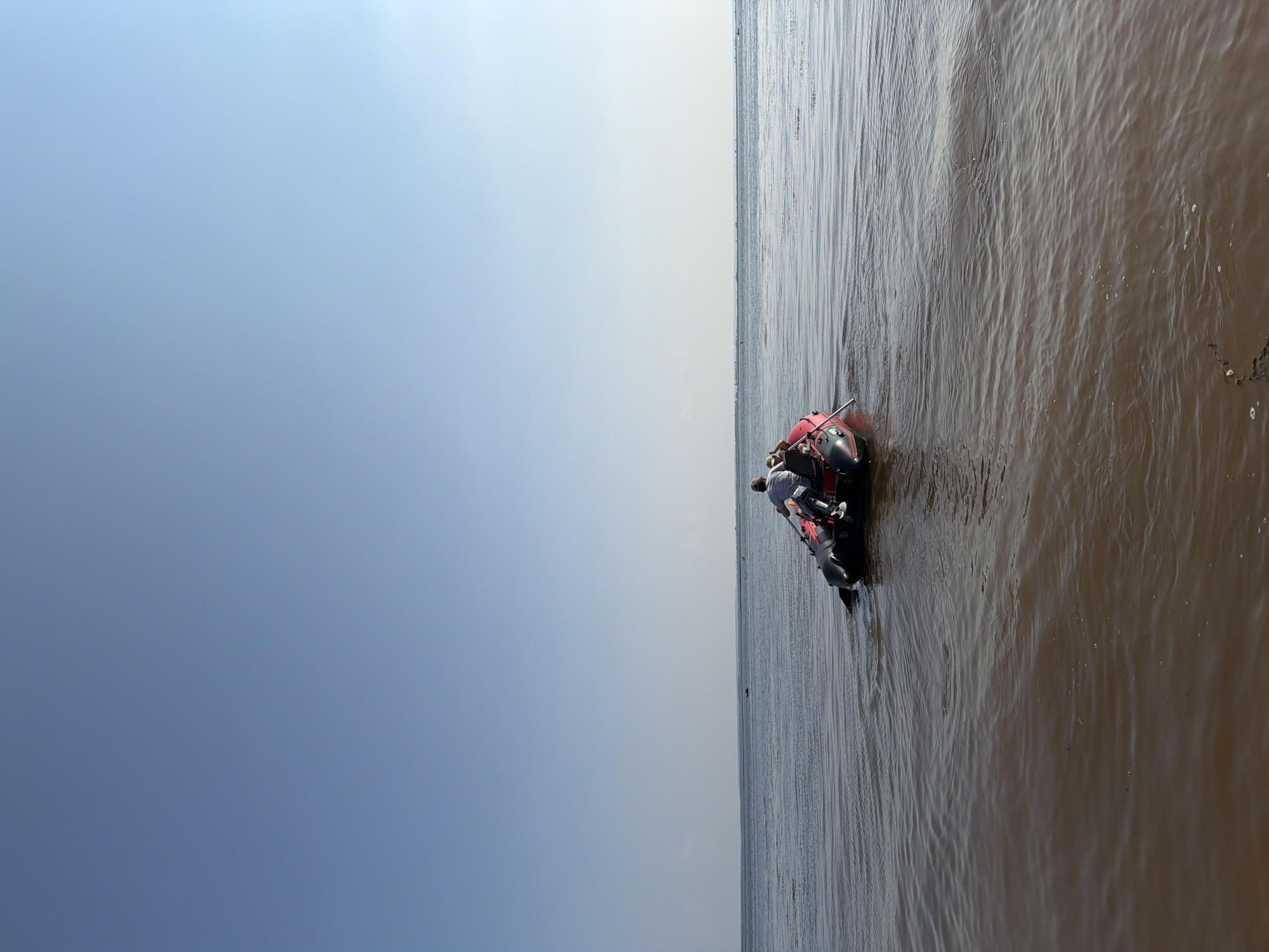 In the city of Krasny Bor, Russia, on a clear day, a red and black inflatable boat with an outboard motor is in the water. Two people are seated in the boat. The person in the front is wearing a light-colored shirt and a cap, and appears to be steering or operating the boat with an oar. The second person is seated behind, facing away from the camera. The water is a murky brown color and appears calm with small ripples. In the distance, a hazy horizon line separates the water from the sky, with a few faint dark objects visible on the horizon that could be other boats or landmasses. The sky is a pale blue and stretches across the upper half of the image.